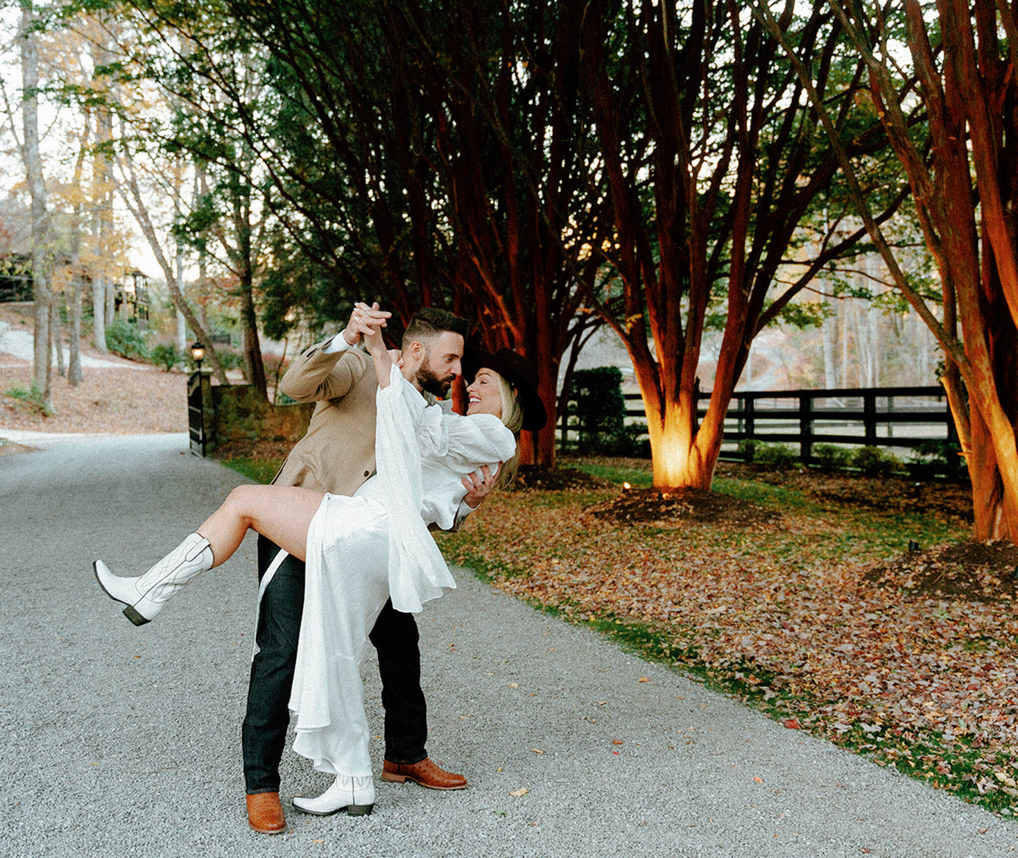 A man in a tan jacket dips and holds a woman in a white dress and cowboy boots on a tree-lined path; both are smiling and surrounded by autumn foliage.