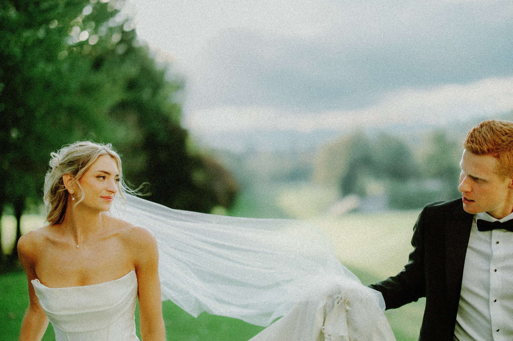 asheville wedding photographer A bride in a white dress and veil smiles at a groom in a black tuxedo as they walk outside on a lush green lawn with trees and a blurred landscape in the background.