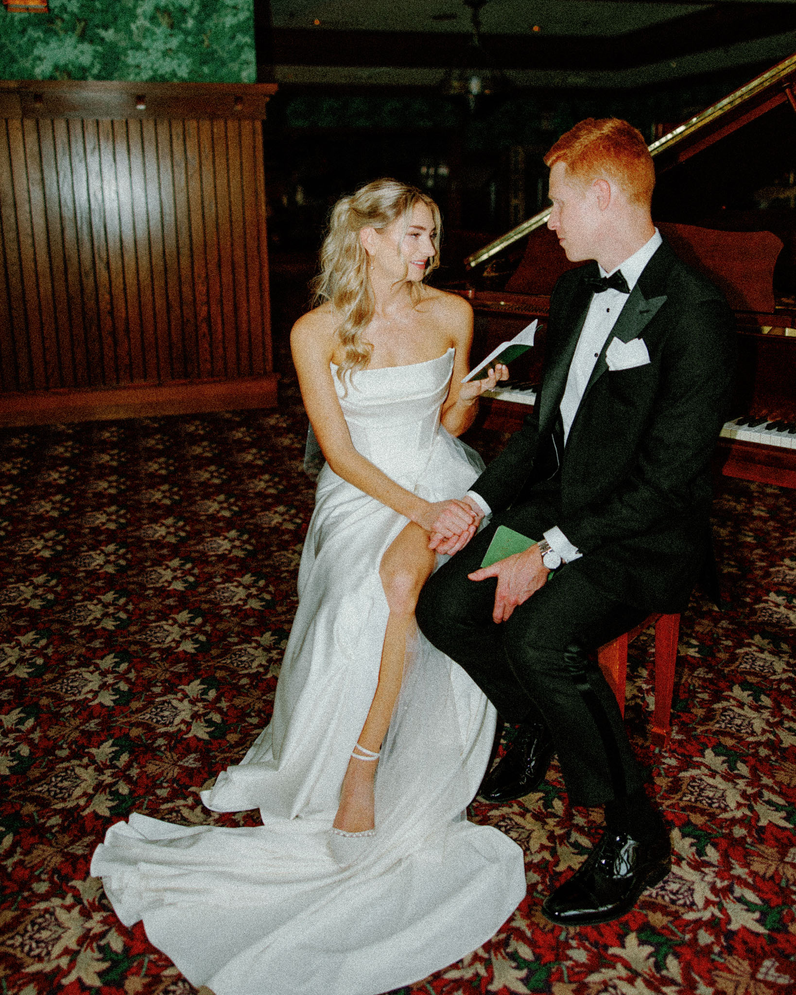 asheville wedding photographer A bride in a white gown sits beside a groom in a black tuxedo on a bench in front of a piano, holding a note and smiling at each other in an elegant, warmly lit room with floral carpet, captured by Grove Park Inn Wedding Photographer on Film.