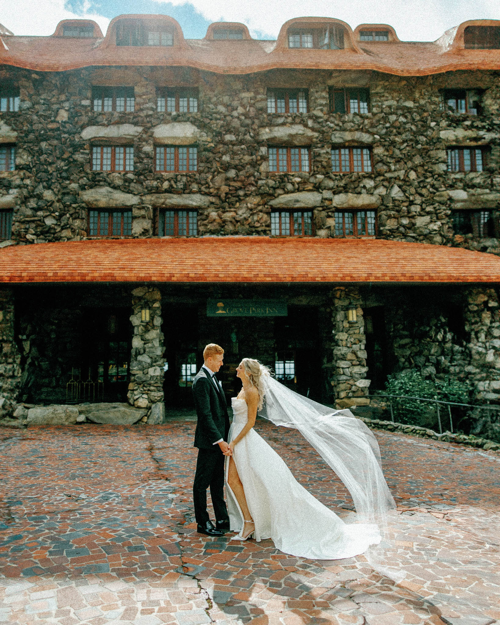 asheville wedding photographer A bride and groom stand holding hands outside a large stone building with a red roof. Captured by a Grove Park Inn Wedding Photographer on film, the bride’s white dress and long veil blow in the wind as the groom stands beside her in a black suit.