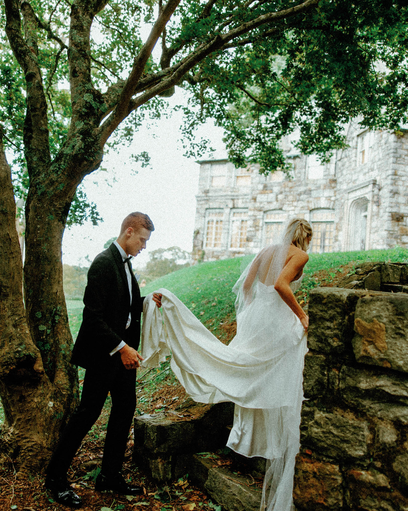asheville wedding photographer A groom in a black suit helps a bride in a white gown and veil up stone steps outdoors, with leafy trees and a large stone building in the background, beautifully captured by a Grove Park Inn Wedding Photographer on Film.