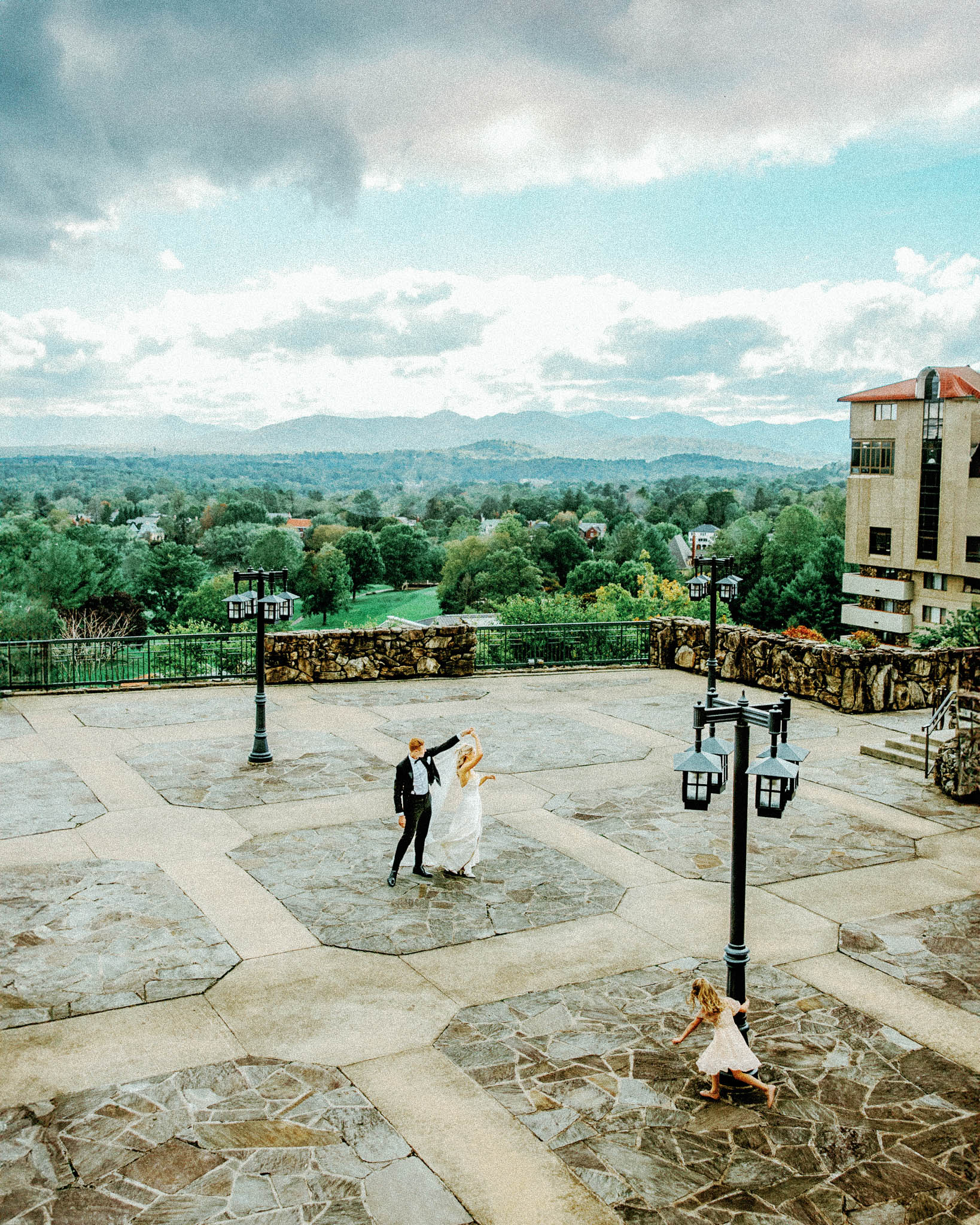 asheville wedding photographer A bride and groom dance on a spacious stone terrace with scenic mountains as their backdrop, while a Grove Park Inn Wedding Photographer on Film kneels to capture the moment under a cloudy sky.