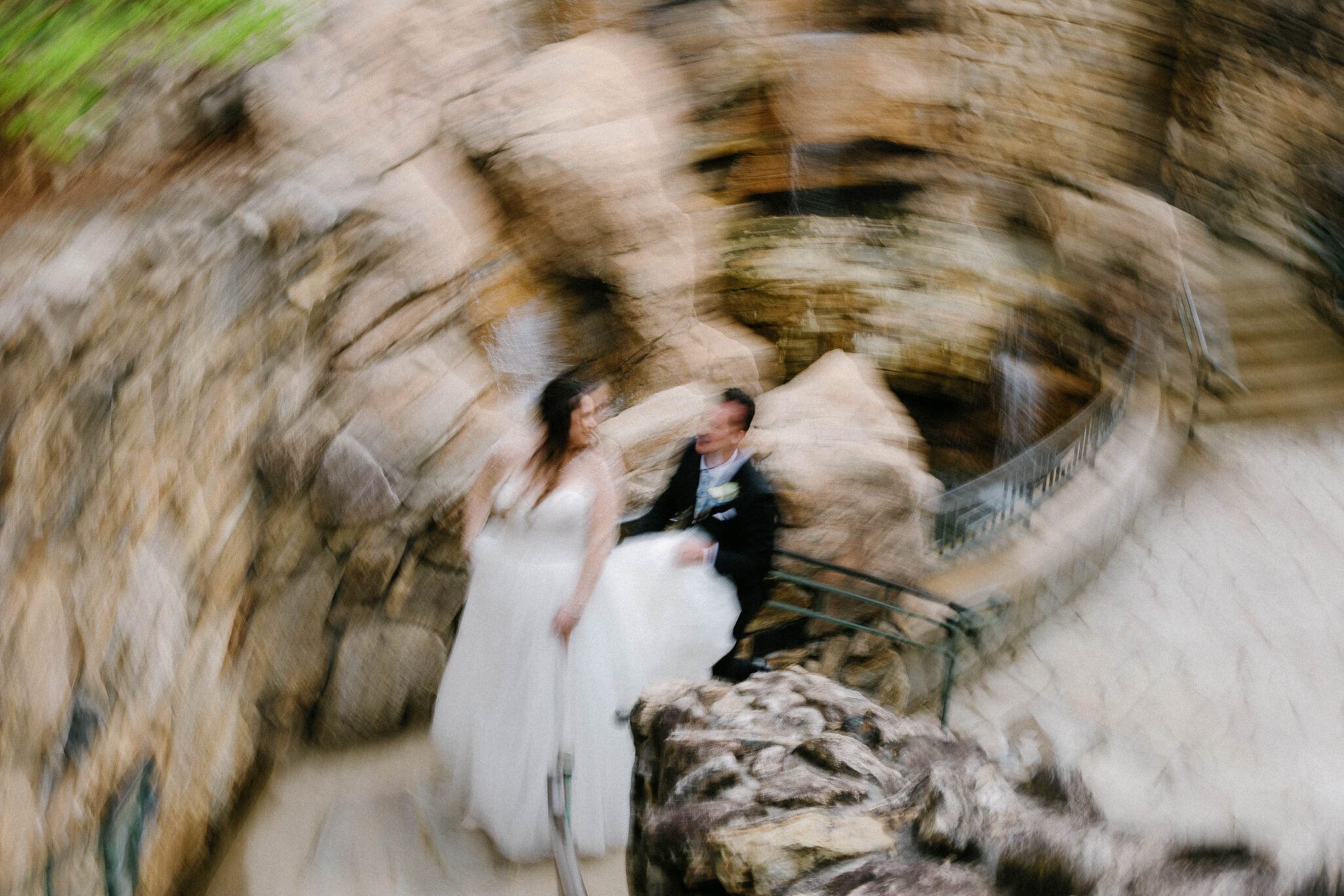 A blurred, swirling photo shows a bride in a white dress and a groom in a suit standing together on a stone staircase surrounded by rocky walls. The motion blur creates a dreamy, abstract effect.