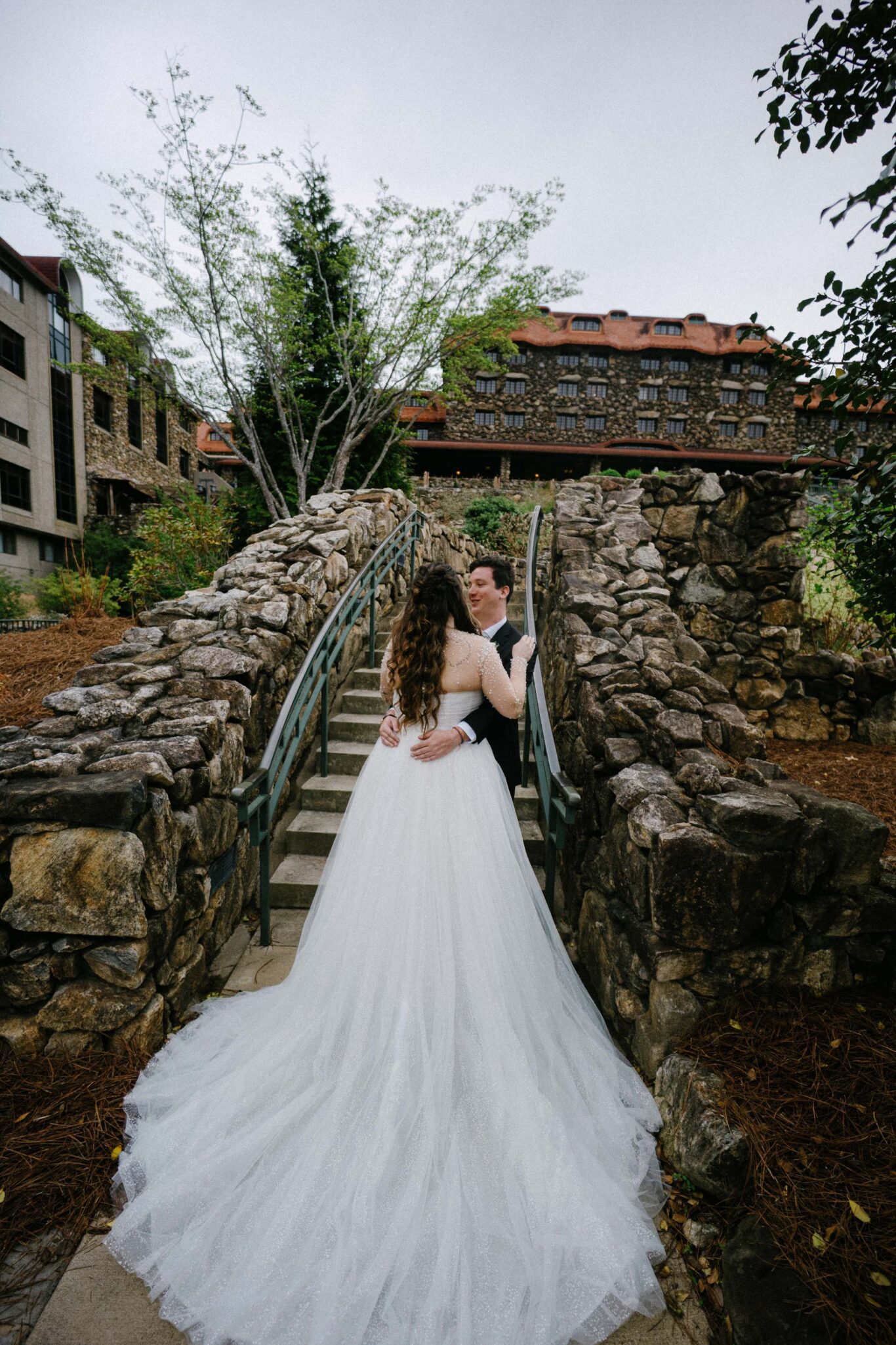 A bride and groom embrace on a stone bridge outside, surrounded by rustic buildings and trees. The bride wears a white gown with a long train, and the groom is in a dark suit, both smiling at each other.
