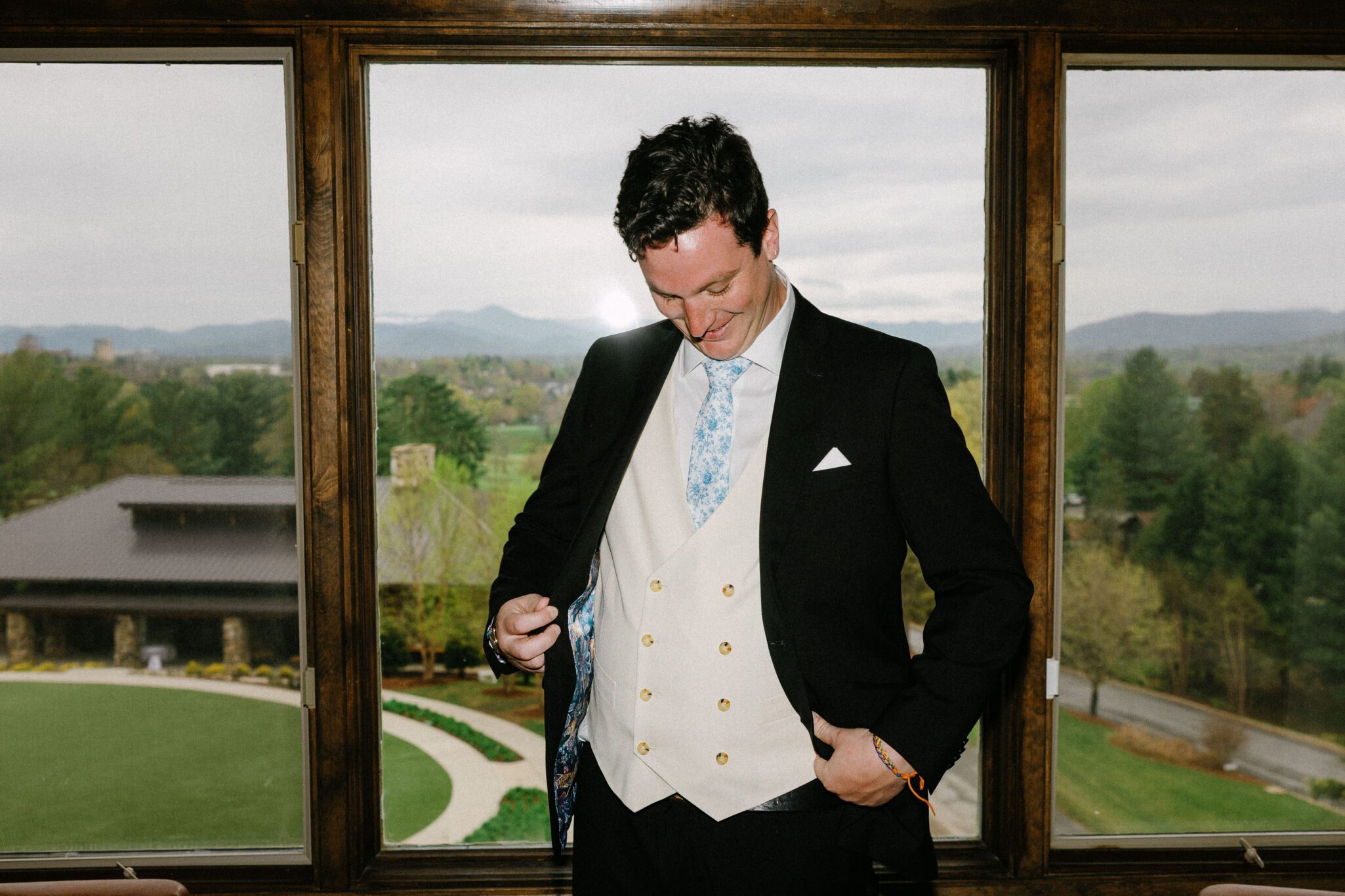A man in a black suit, light blue tie, and cream-colored vest smiles while looking down and adjusting his suit in front of a large window with a scenic view of greenery and distant mountains.