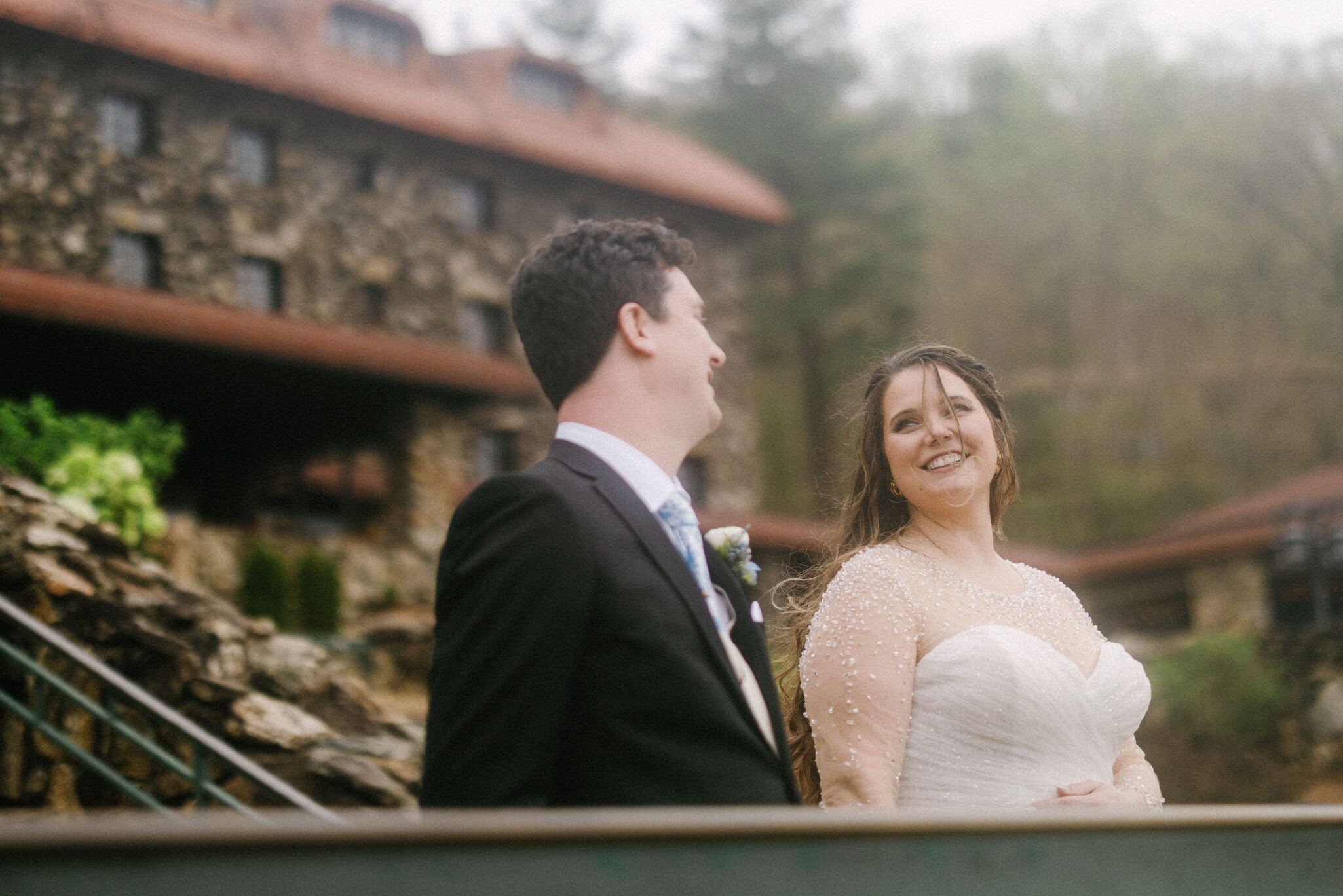A bride and groom stand outdoors, smiling at each other in front of a rustic stone building with a red roof and surrounded by greenery. The bride wears a white dress and the groom is in a black suit with a tie.