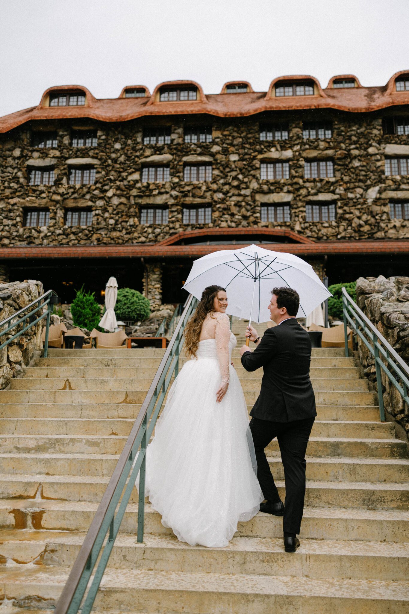A bride and groom walk up stone steps outside a large rustic building. The groom holds a white umbrella over them as they smile at each other, with the bride looking back over her shoulder.
