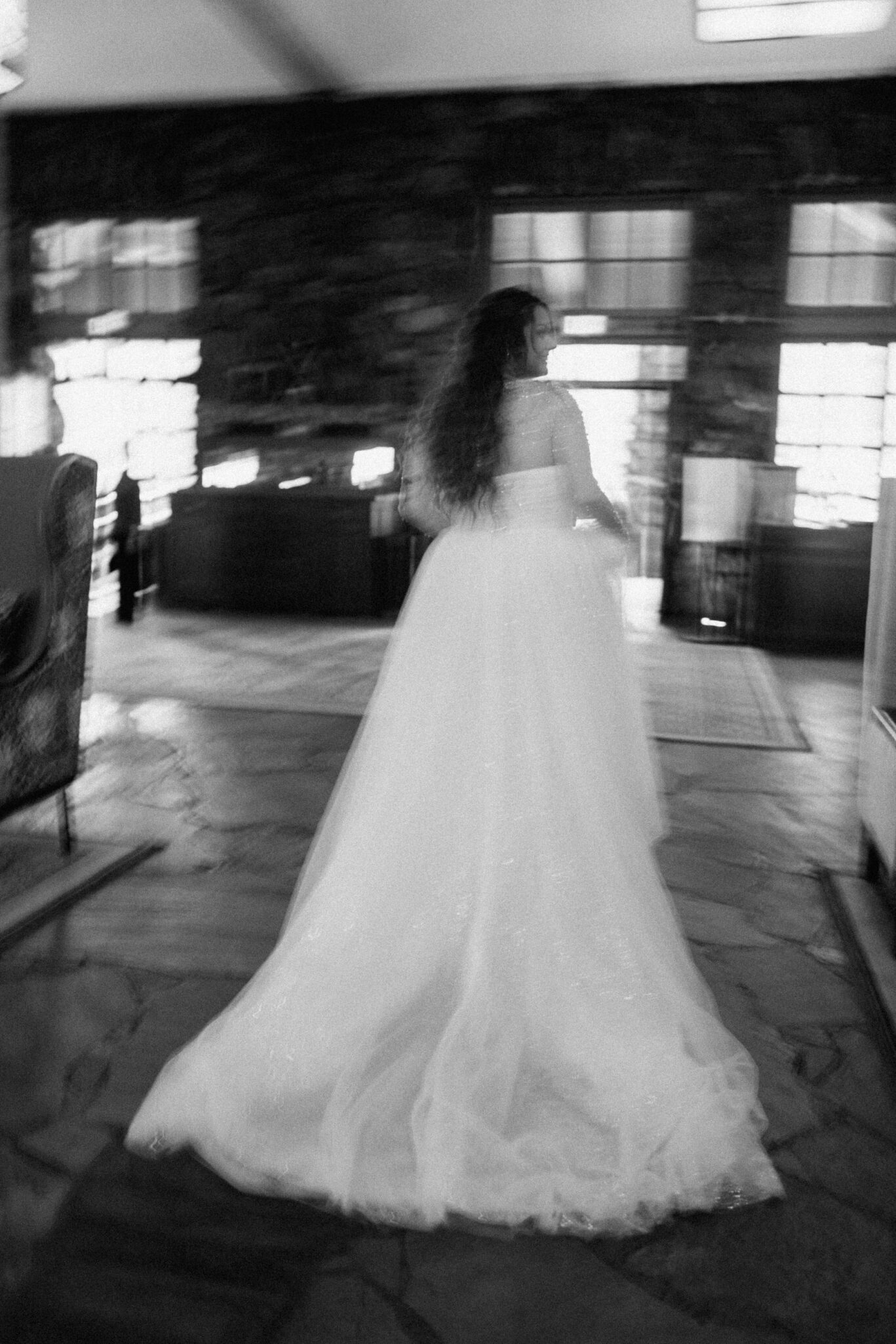 A black-and-white photo of a woman in a long, flowing bridal gown walking indoors, away from the camera, in a spacious room with stone walls and large windows.