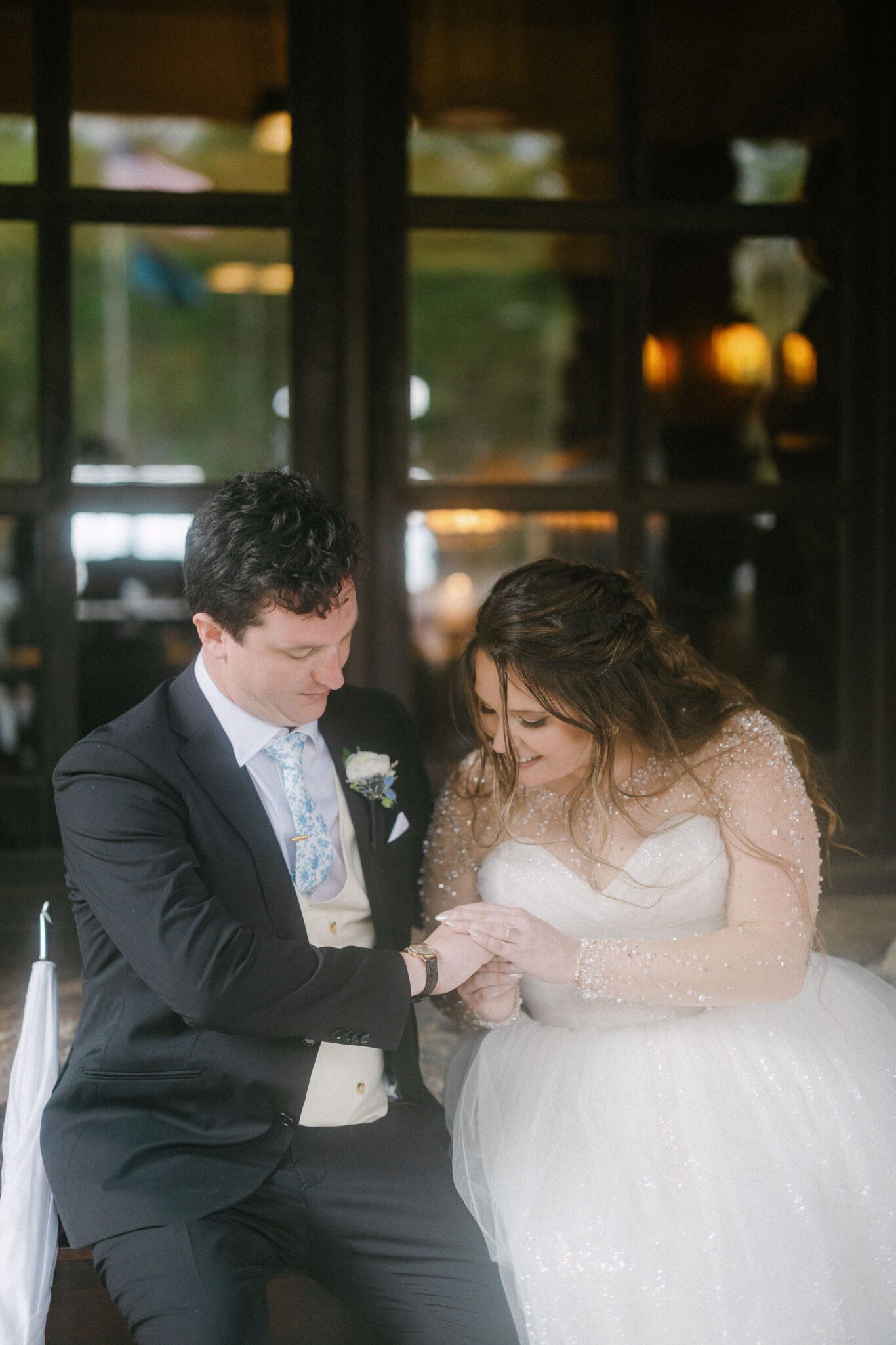 A bride in a white gown and groom in a suit sit closely together, smiling as the bride gently holds the groom’s hand—an intimate, joyful moment beautifully captured by The Omni Grove Park Inn wedding photographer amid softly blurred lights and greenery.