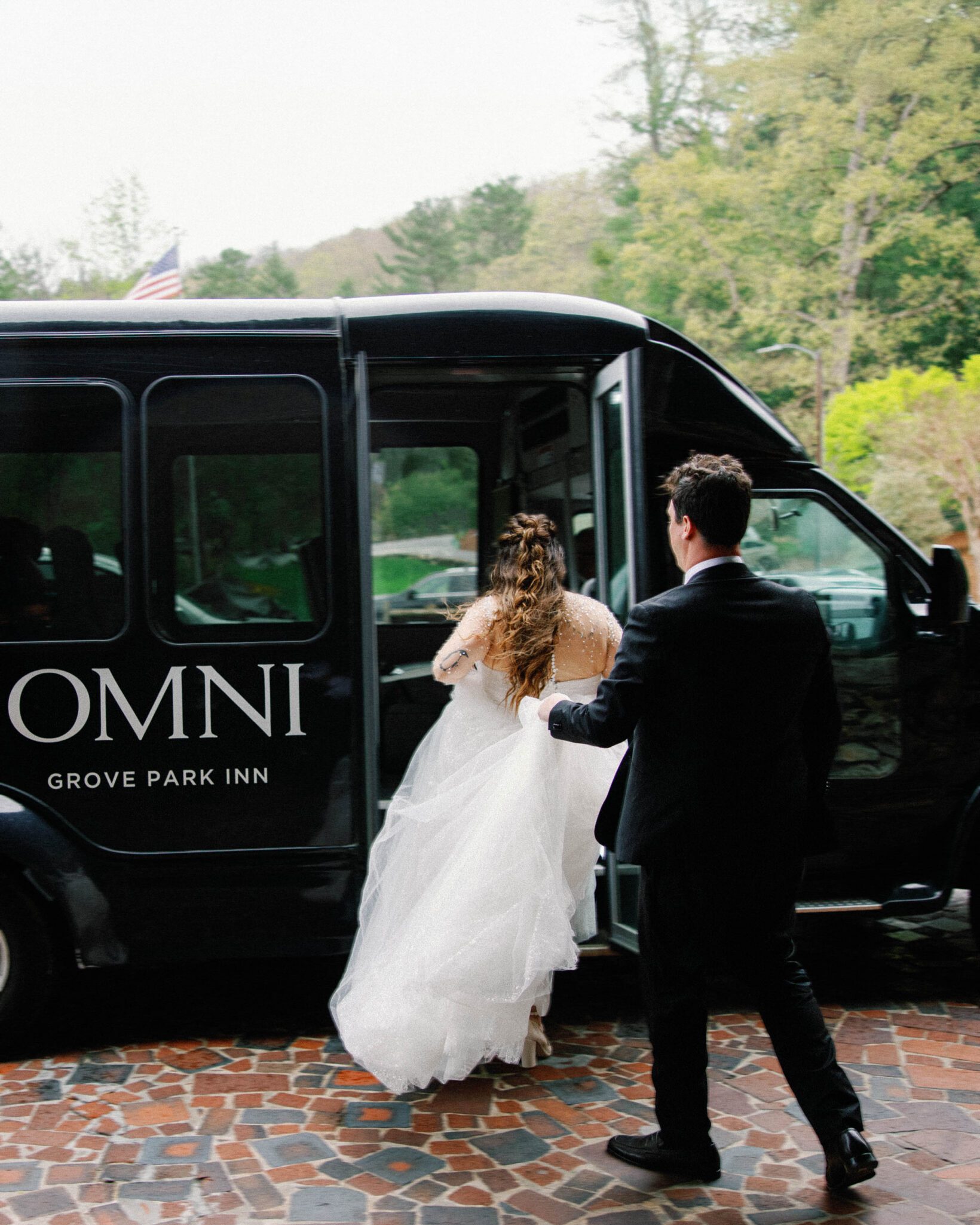 A bride in a white dress steps into a black Omni Grove Park Inn shuttle bus as a man in a suit stands nearby, gently holding her arm. The scene takes place outdoors on a stone path with greenery in the background.