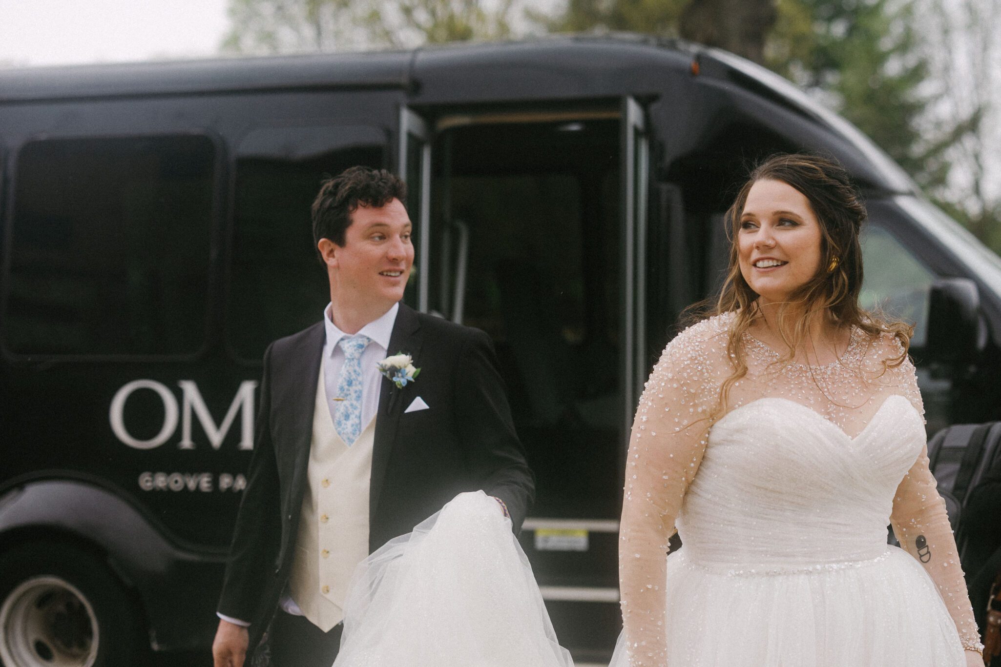 A bride in a white gown with pearl details and a groom in a black suit with a cream vest walk outside, smiling, in front of a black van with the door open.