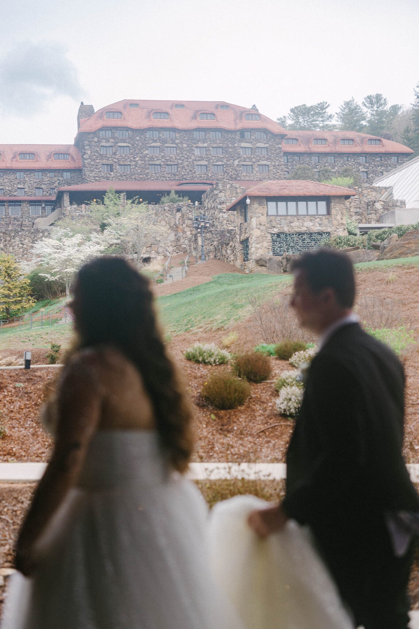 A bride and groom, both out of focus, walk outdoors with a large stone hotel or lodge in the background amid trees and landscaped grounds.