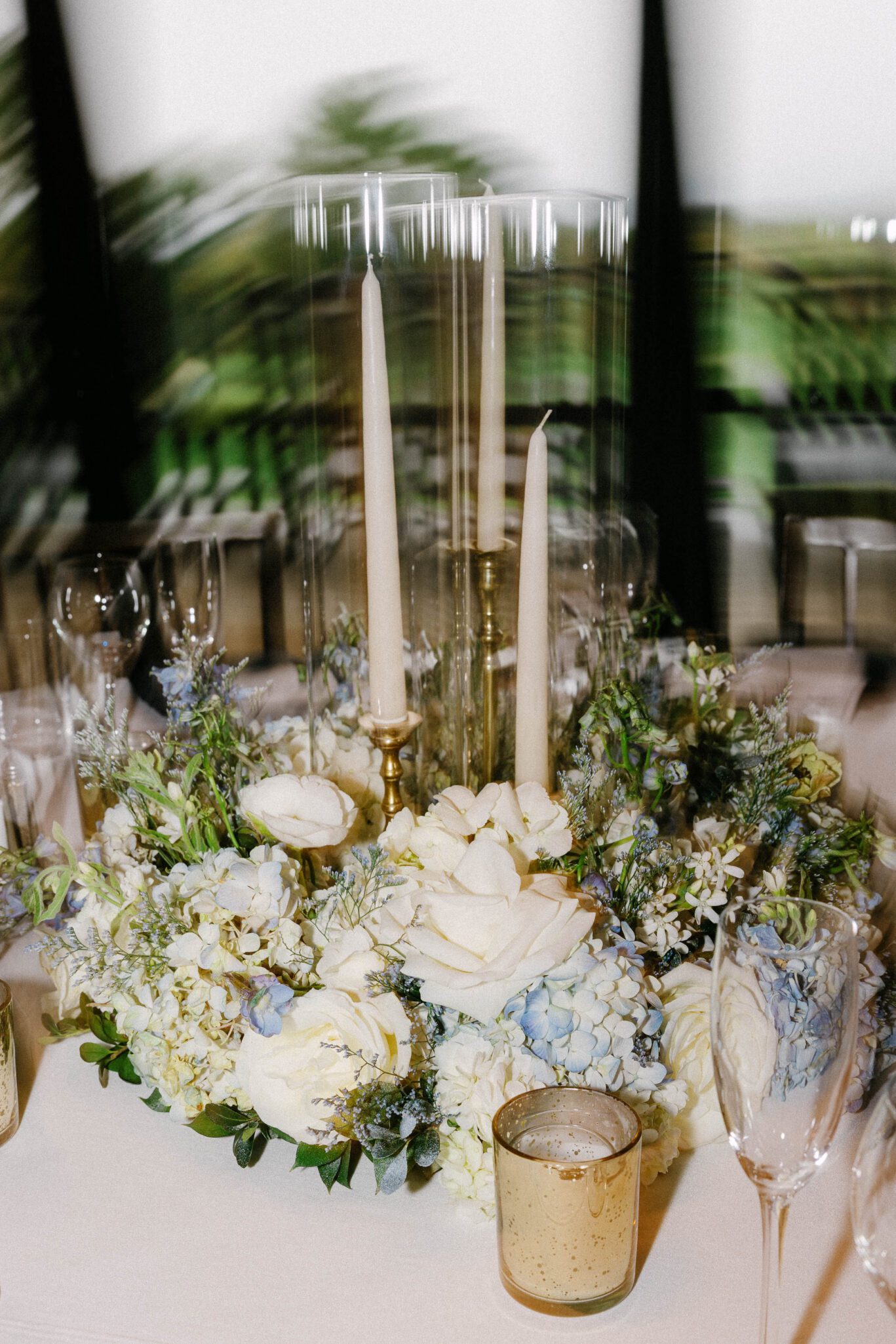 A table centerpiece featuring two tall white taper candles in glass holders, surrounded by white and pale blue flowers, greenery, and gold votive candle holders on a white tablecloth.