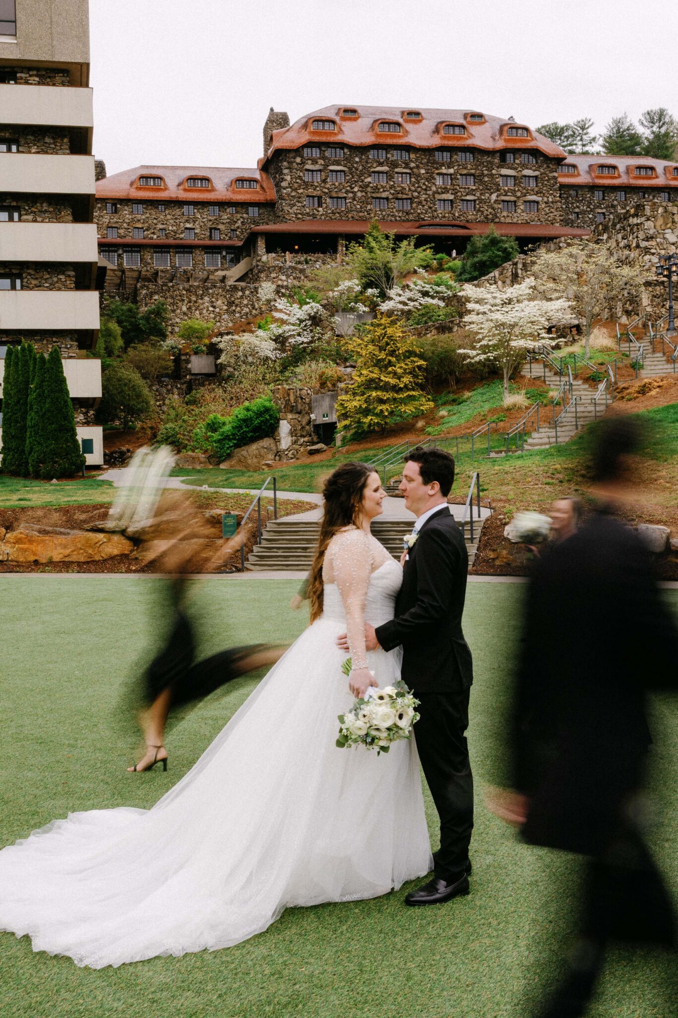 A bride and groom stand facing each other on a lawn, the bride holding a bouquet. People in black clothing walk by in blurred motion, and a large historic building and greenery are in the background.