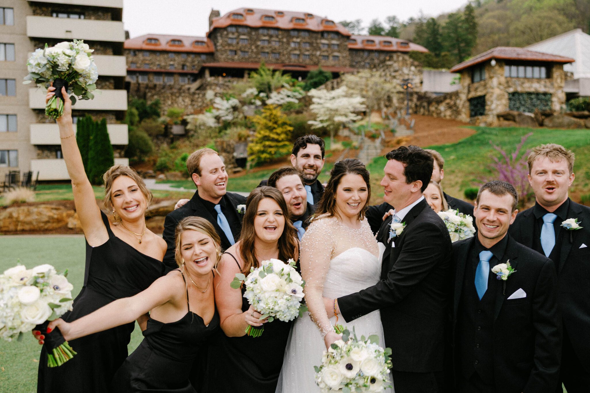 A joyful bride and groom stand outdoors with their wedding party, all smiling and posing together. The bridesmaids wear black dresses, and the groomsmen wear black suits with blue ties. A rustic building and greenery are in the background.