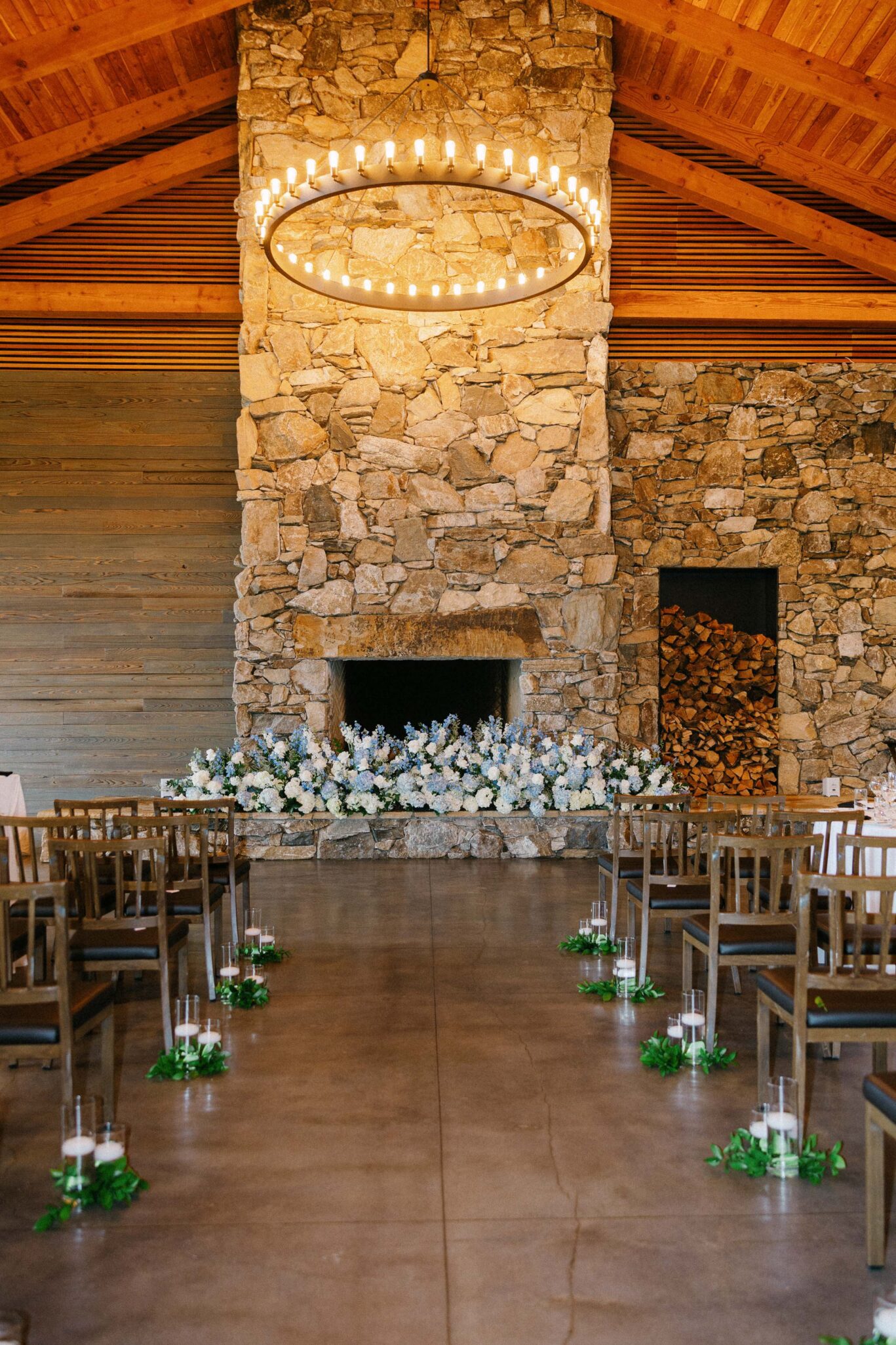 A rustic indoor wedding ceremony setup at The Omni Grove Park Inn, featuring a stone fireplace with a flower arrangement, wooden chairs lined with greenery and candles, and a large circular chandelier hanging from a wooden ceiling—perfect for any wedding photographer.