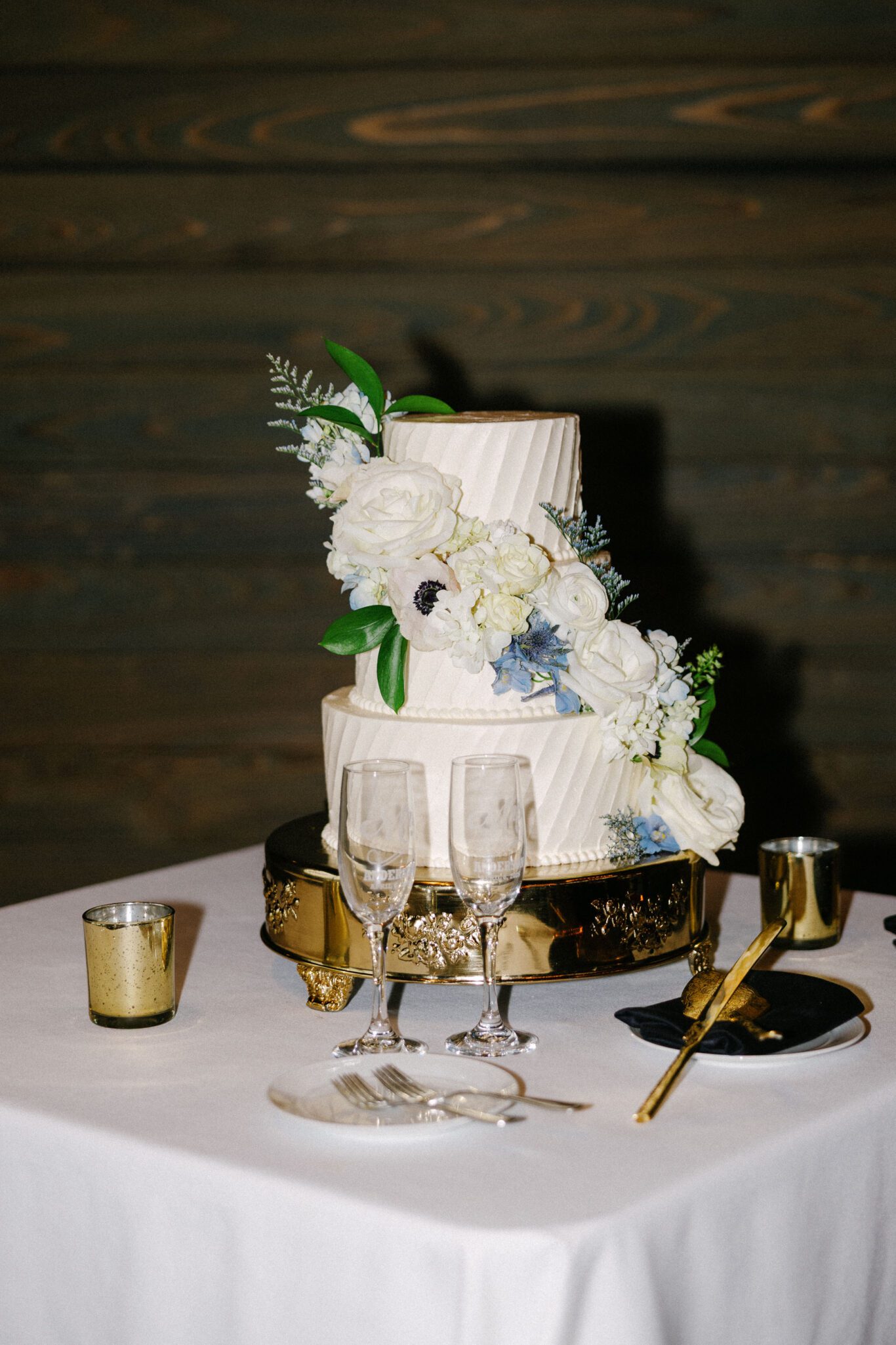A two-tiered white wedding cake decorated with white and blue flowers sits on a gold stand. The cake is on a white table with two champagne flutes, gold utensils, and gold candle holders.