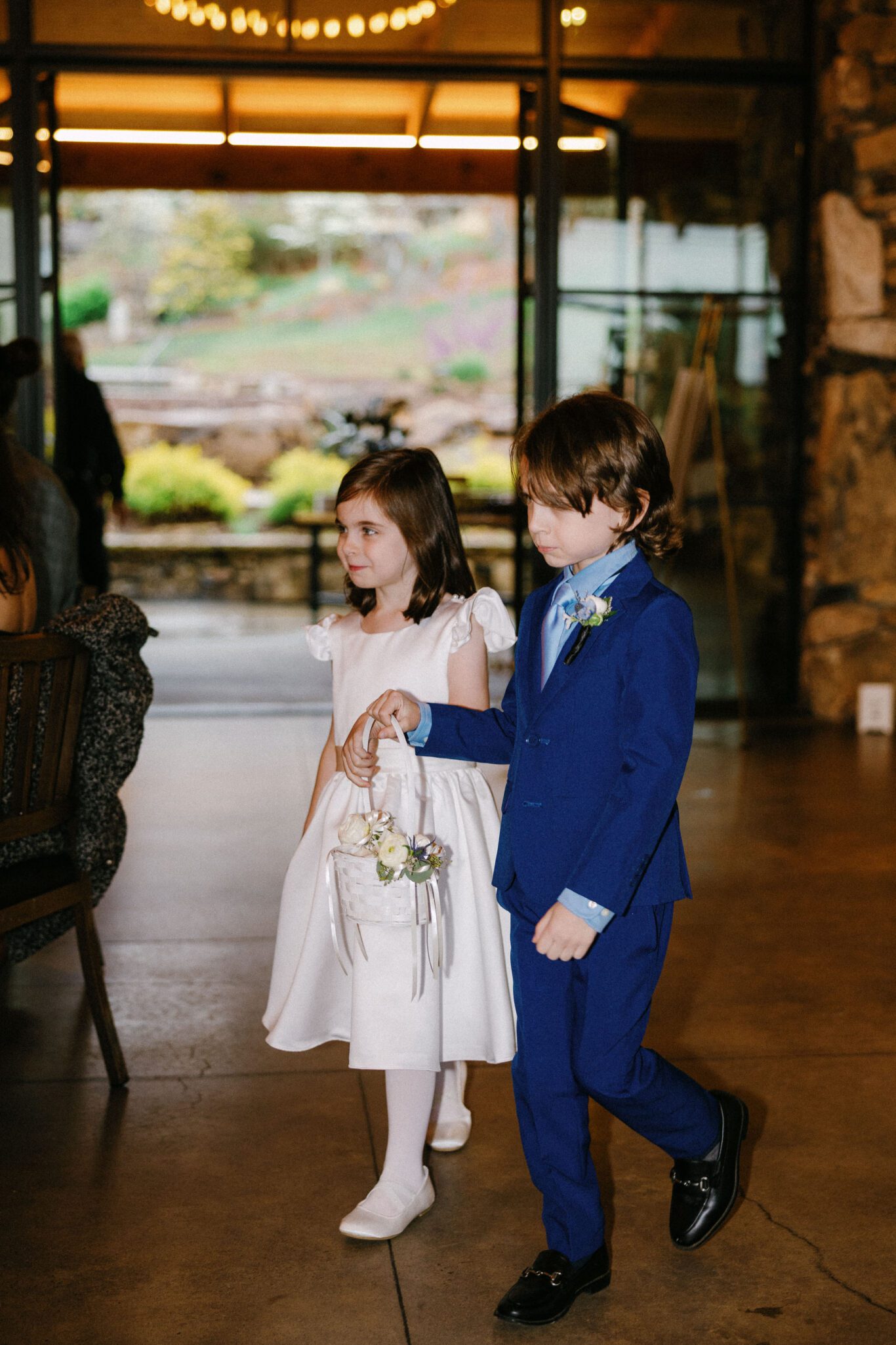A young girl in a white dress and a young boy in a blue suit walk together indoors; the girl carries a basket of flowers. They appear to be participating in a formal event, possibly a wedding.