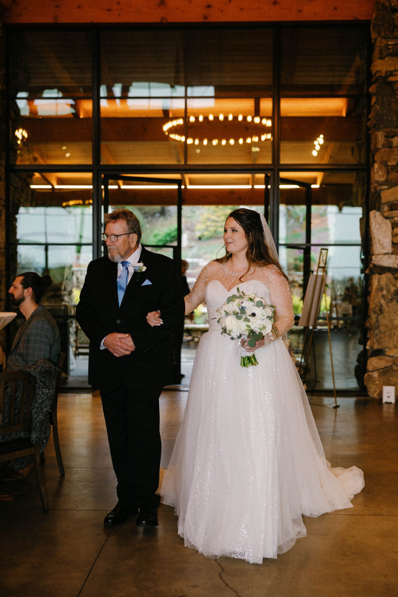 A bride in a white, sparkling gown holds a bouquet of white flowers and walks arm-in-arm with an older man in a black suit, inside a warmly lit venue with stone walls and large windows.