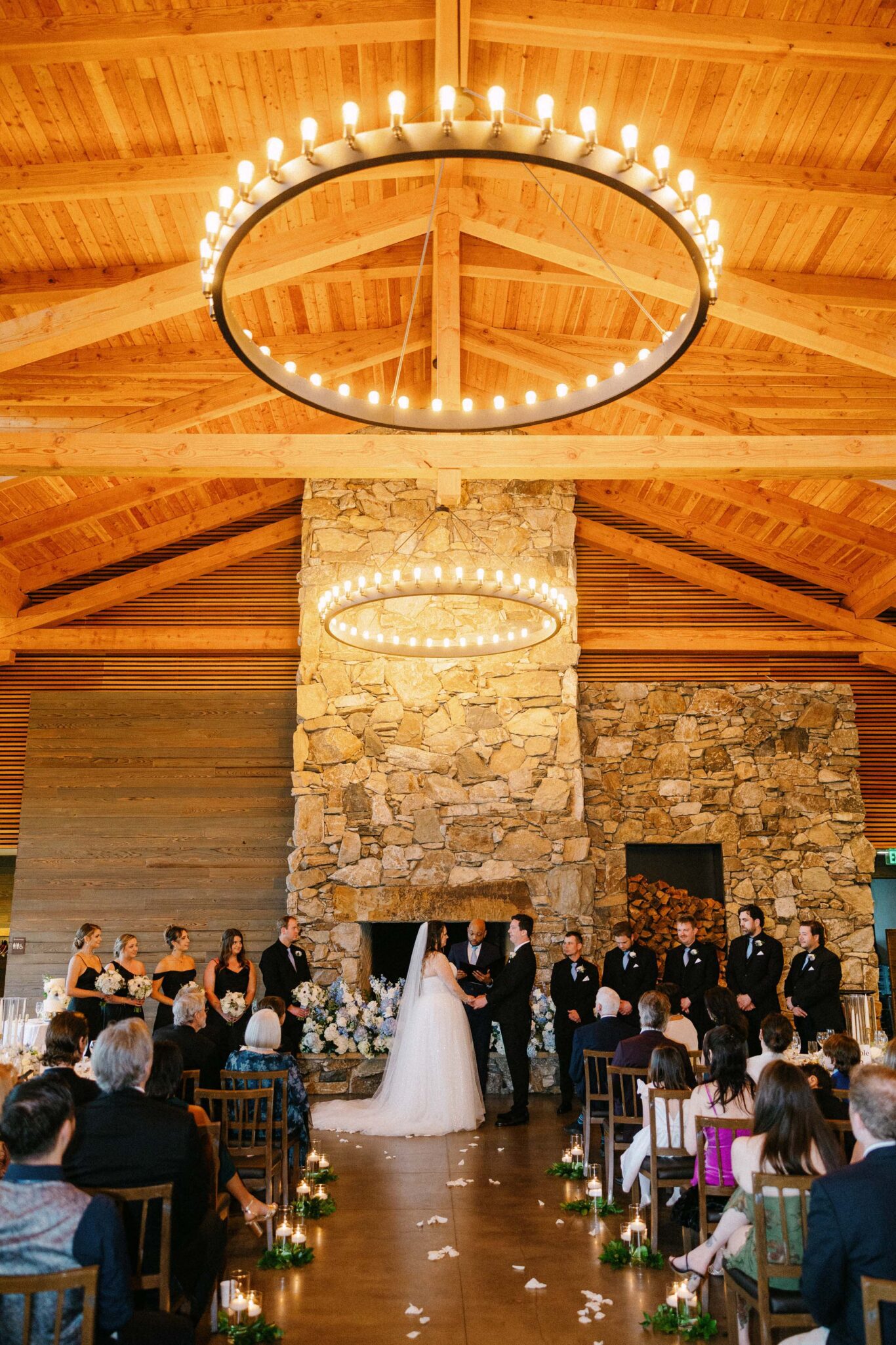 A bride and groom stand at the altar in front of a large stone fireplace, surrounded by their wedding party, under two large circular chandeliers in a wooden hall filled with seated guests.
