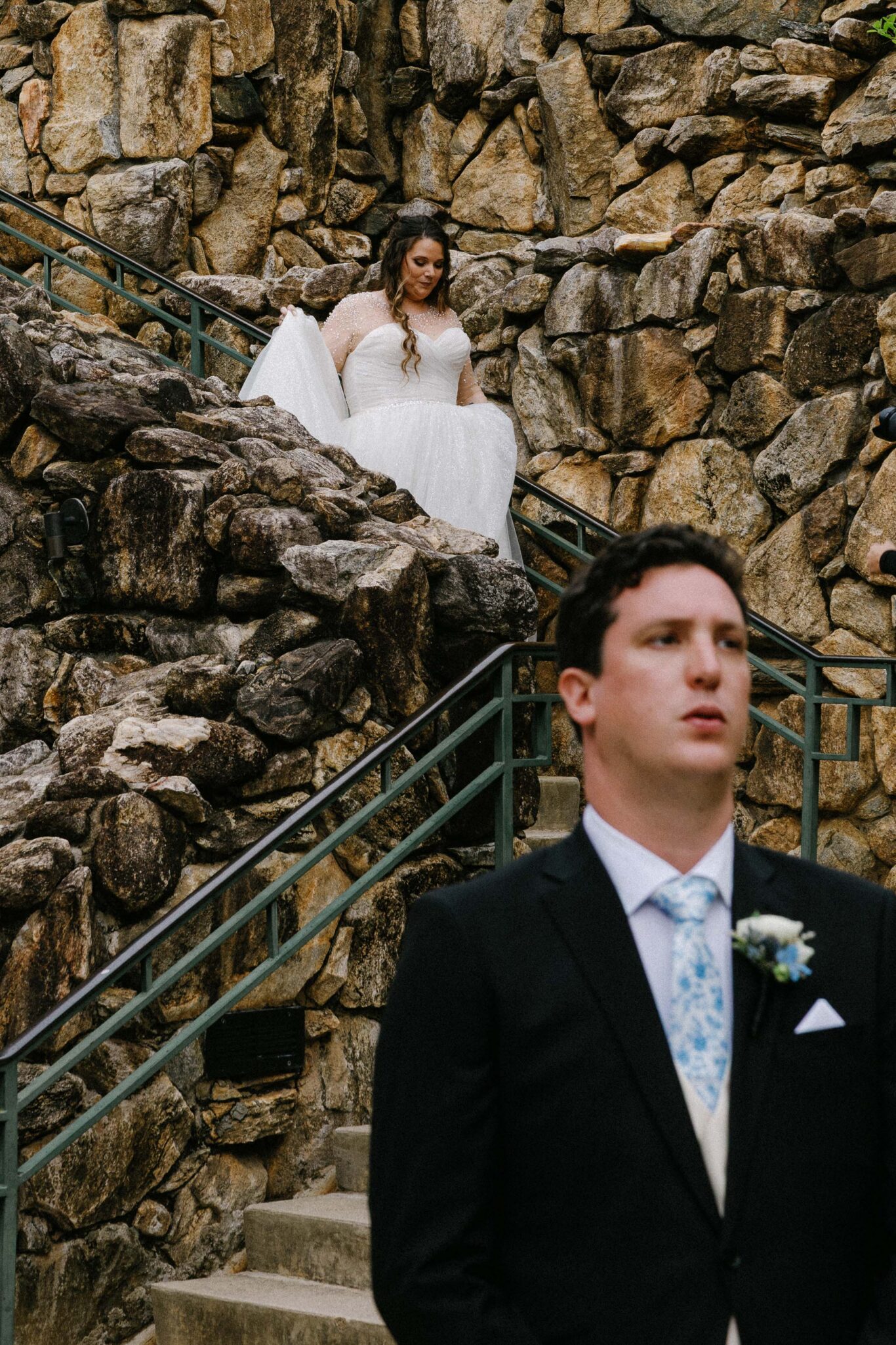 A bride in a white dress descends stone steps, holding her gown, while a groom in a black suit stands in the foreground with a serious expression, both outdoors against a rocky backdrop.
