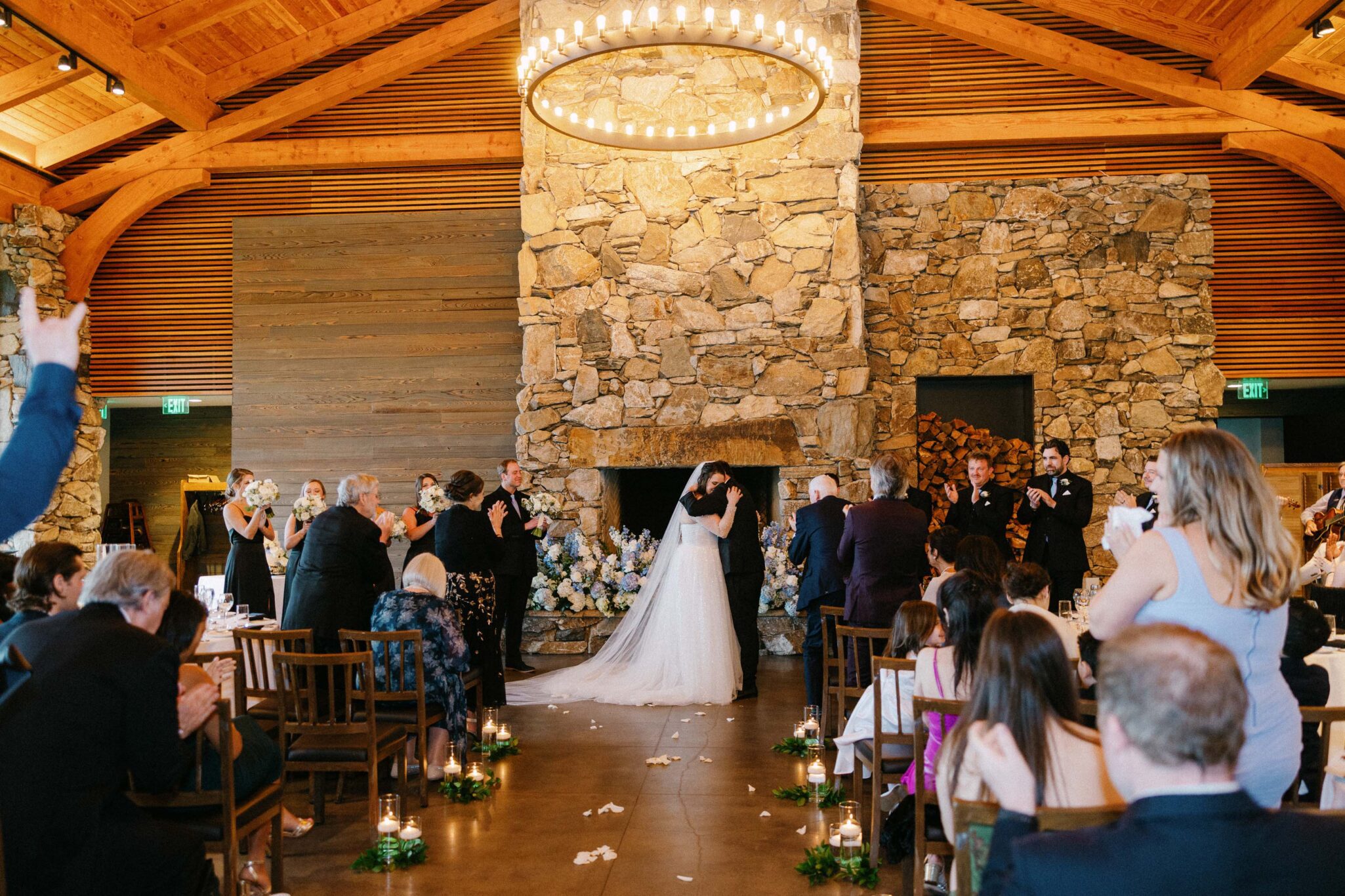 A bride and groom share their first kiss at the altar in a rustic venue with stone walls, surrounded by seated and standing guests clapping and cheering. Candles and greenery line the aisle. A large round chandelier hangs above.