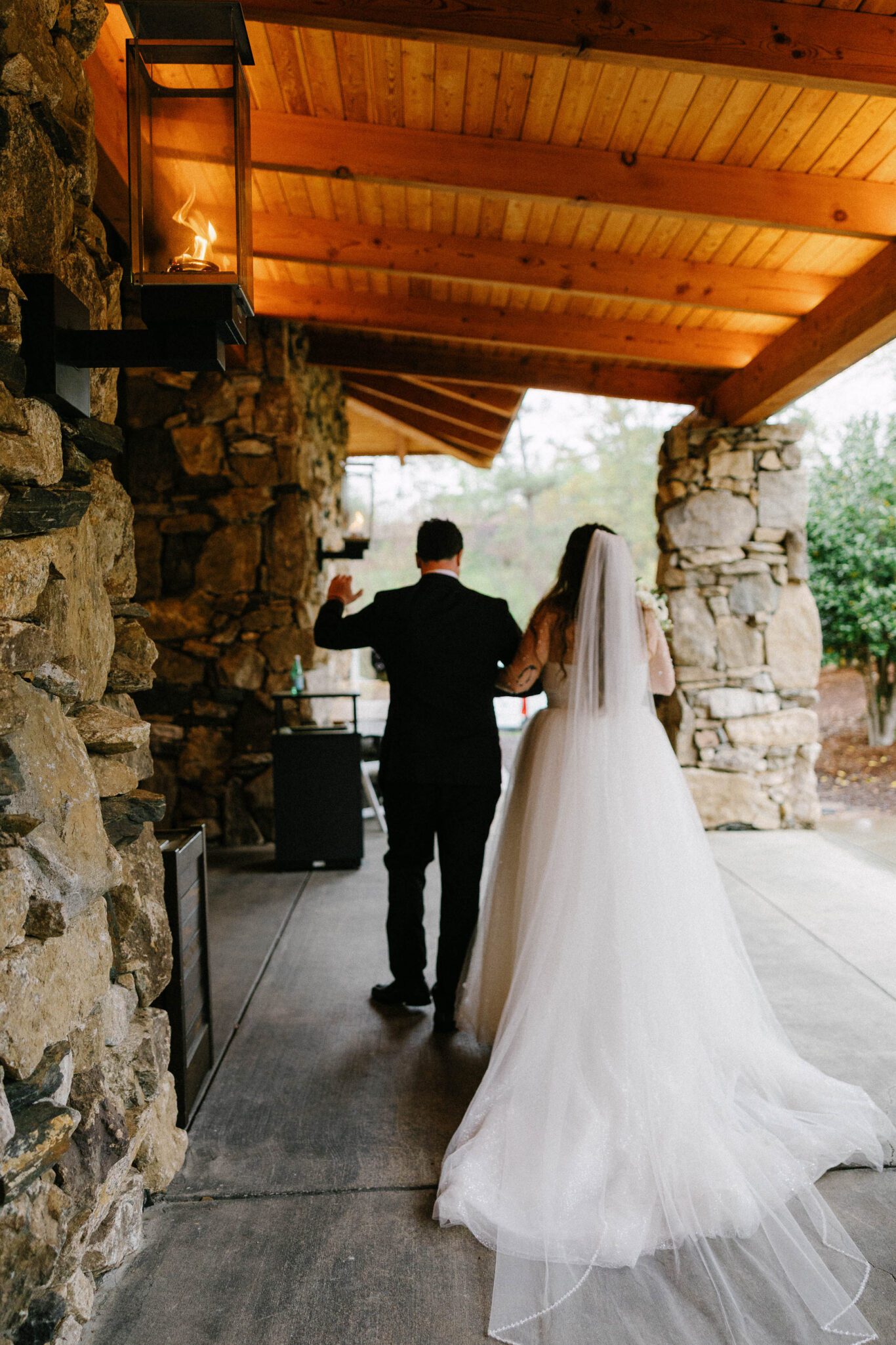 A bride in a white gown and veil walks arm in arm with a groom in a black suit under a rustic stone and wood pavilion, their backs to the camera as they celebrate together.