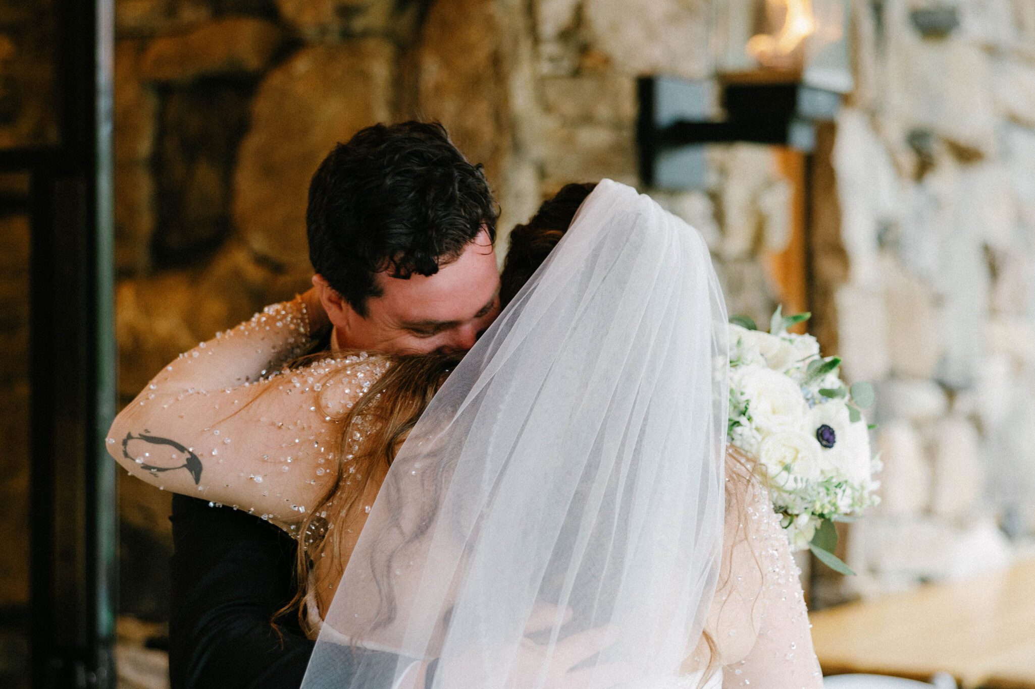 A bride in a white veil hugs a groom closely in front of a stone wall, her bouquet of white flowers visible as they share an intimate moment on their wedding day.