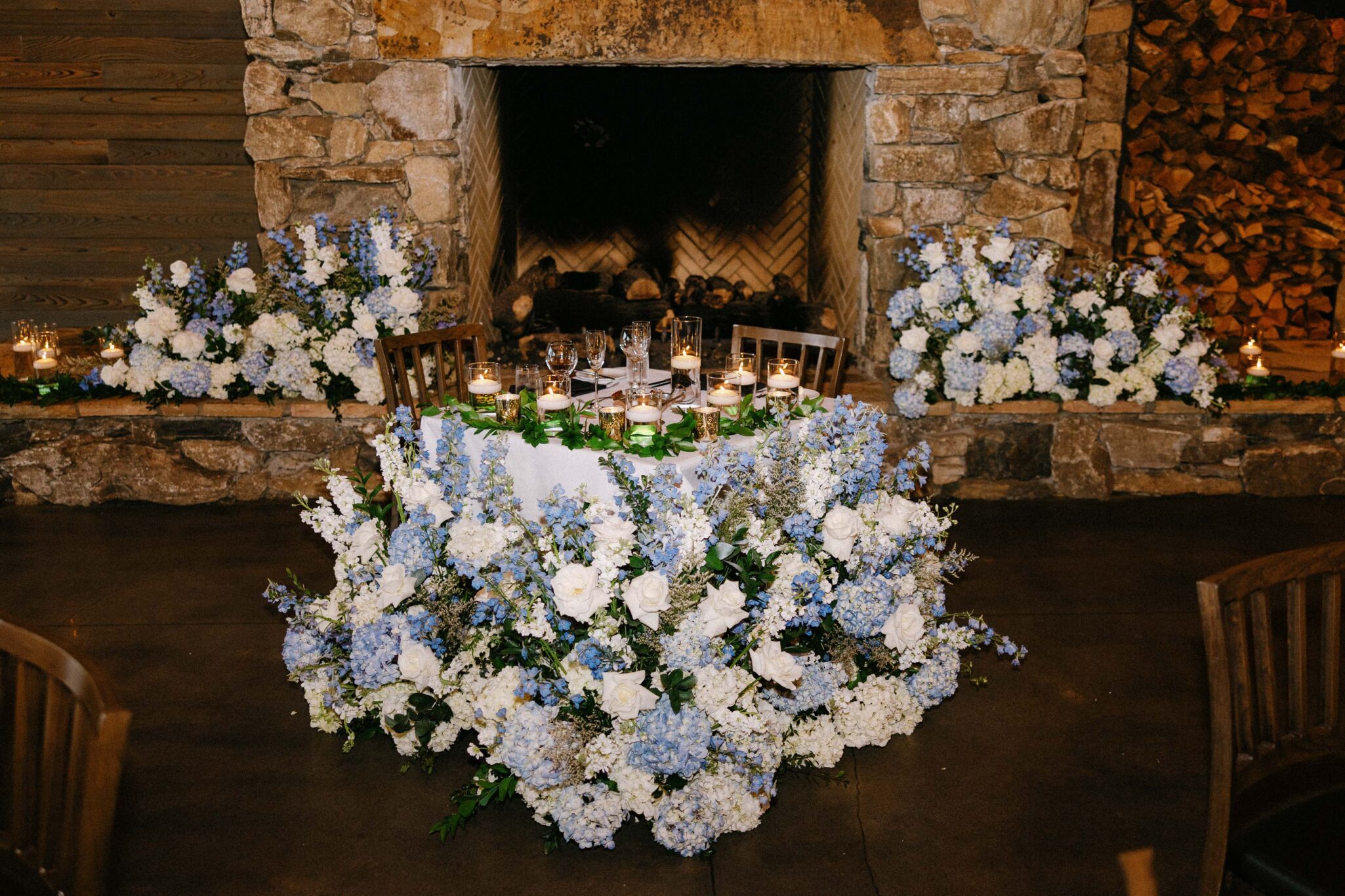 A round table covered in white and blue flowers and surrounded by candles sits in front of a stone fireplace, creating an elegant and romantic setting.