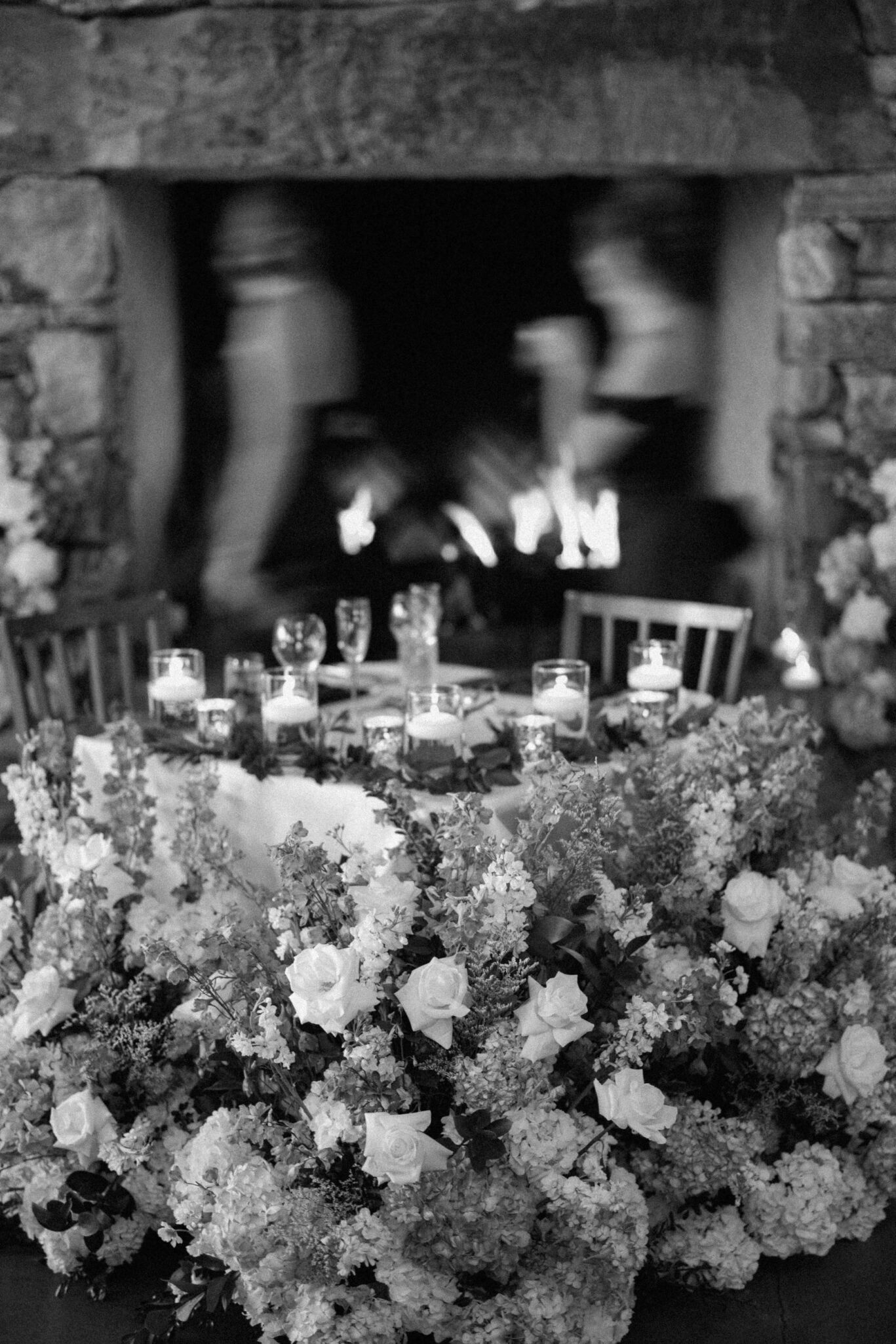 A black and white photo of an elegantly set table with candles and glassware, surrounded by abundant floral arrangements. In the background, two blurred figures stand near a stone fireplace with a lit fire.
