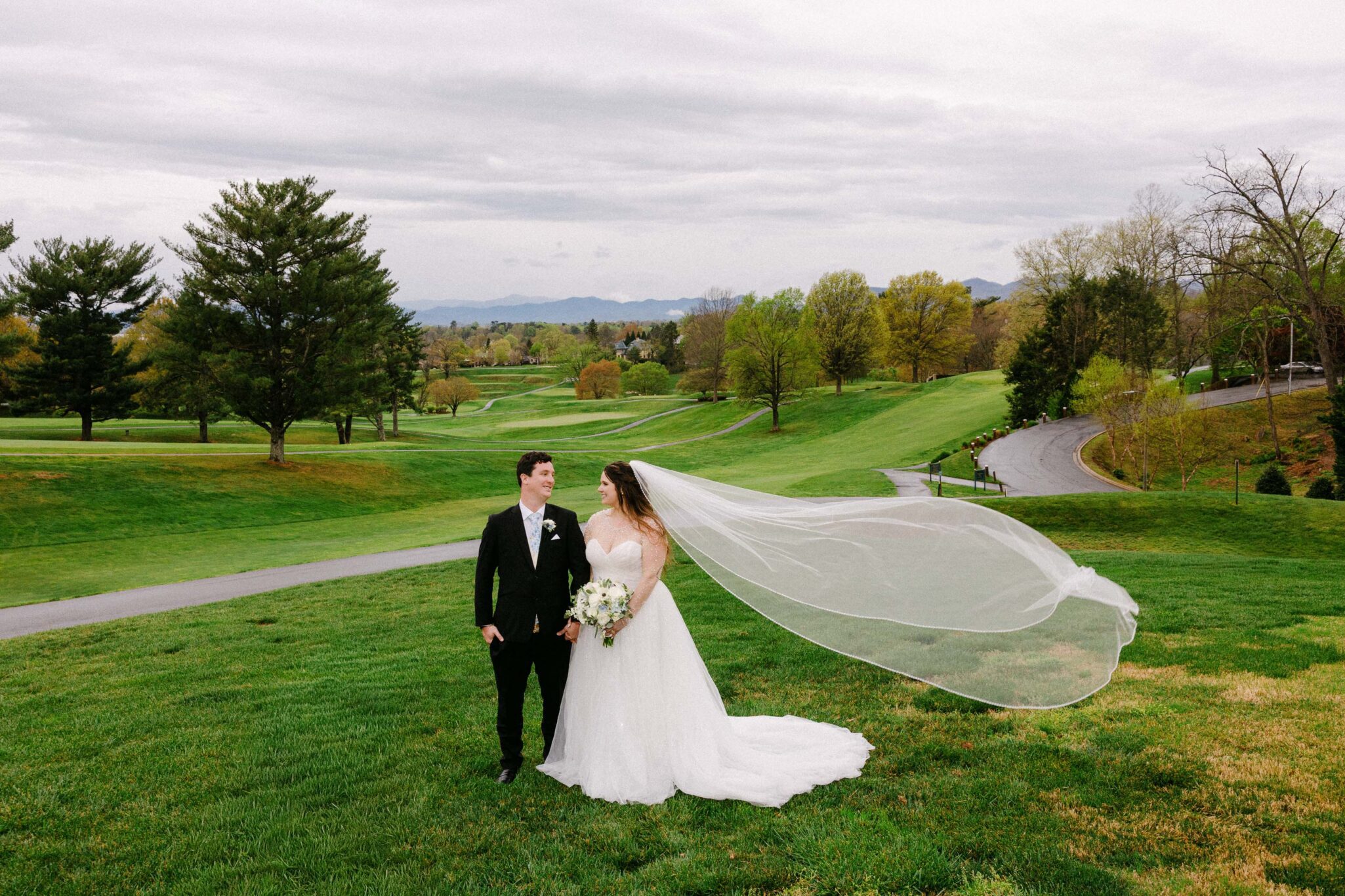 A bride and groom stand on a grassy hill, smiles bright as the wind lifts her veil. Captured by The Omni Grove Park Inn wedding photographer, they are surrounded by green trees, golf paths, and rolling hills beneath a cloudy sky.