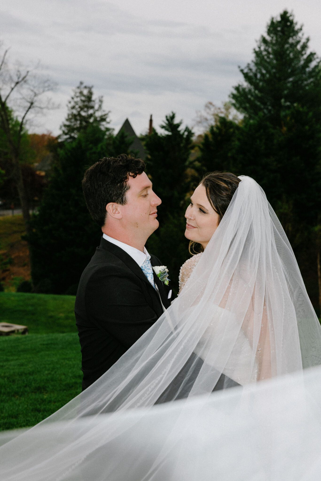 A bride and groom embrace outdoors, gazing at each other lovingly. The bride’s long white veil flows around them, and they are surrounded by green trees and grass under an overcast sky.