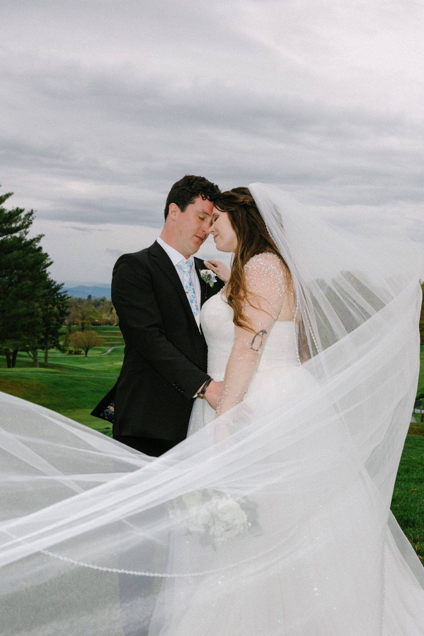 A bride and groom stand closely together outdoors, heads touching lovingly. The brides long veil flows dramatically in the wind, and the groom wears a black suit. Green grass and cloudy sky form the background.