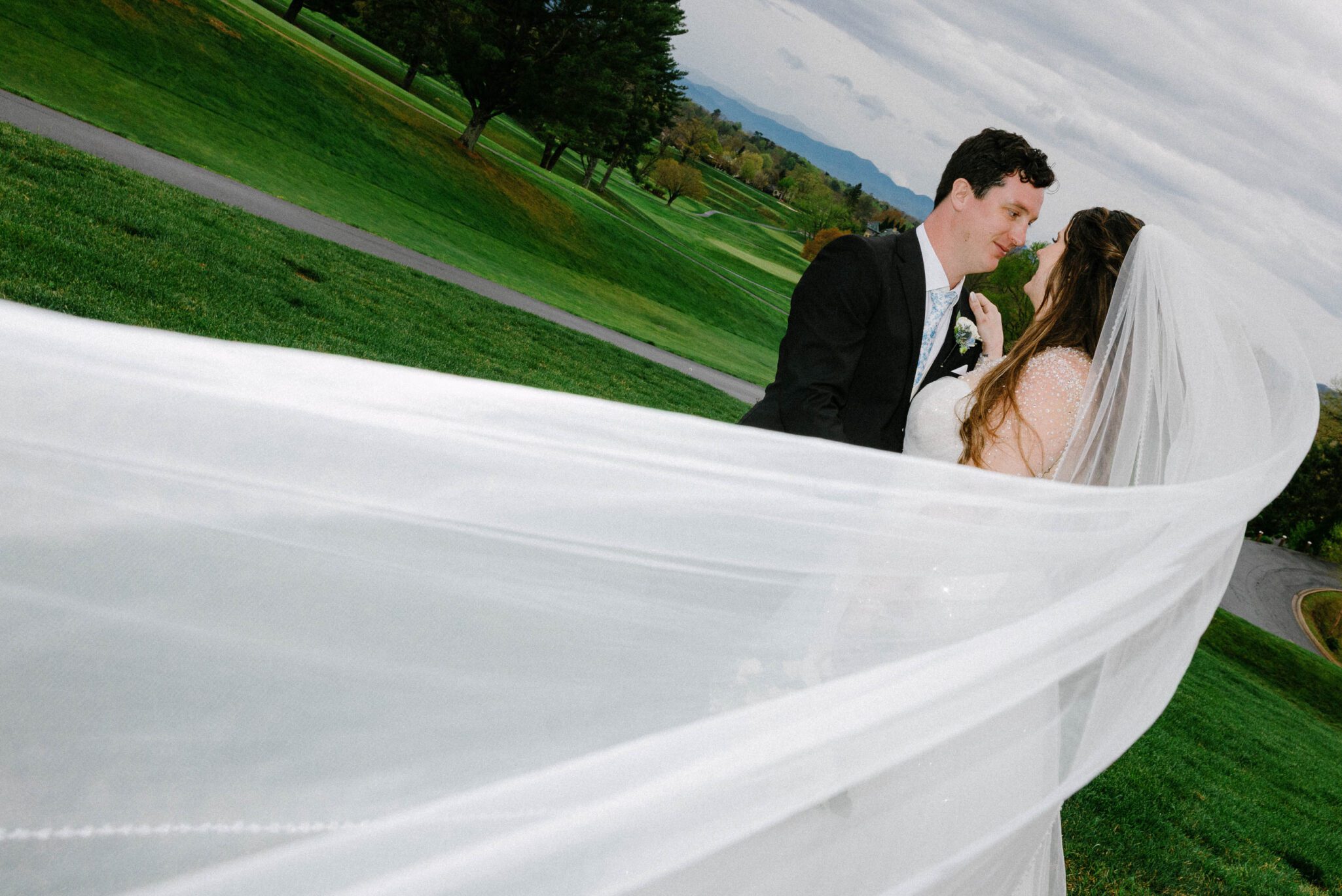 A bride and groom stand outdoors on a green lawn, gazing at each other lovingly. The brides long veil flows toward the camera, creating a sweeping effect. The background shows a cloudy sky and distant trees.