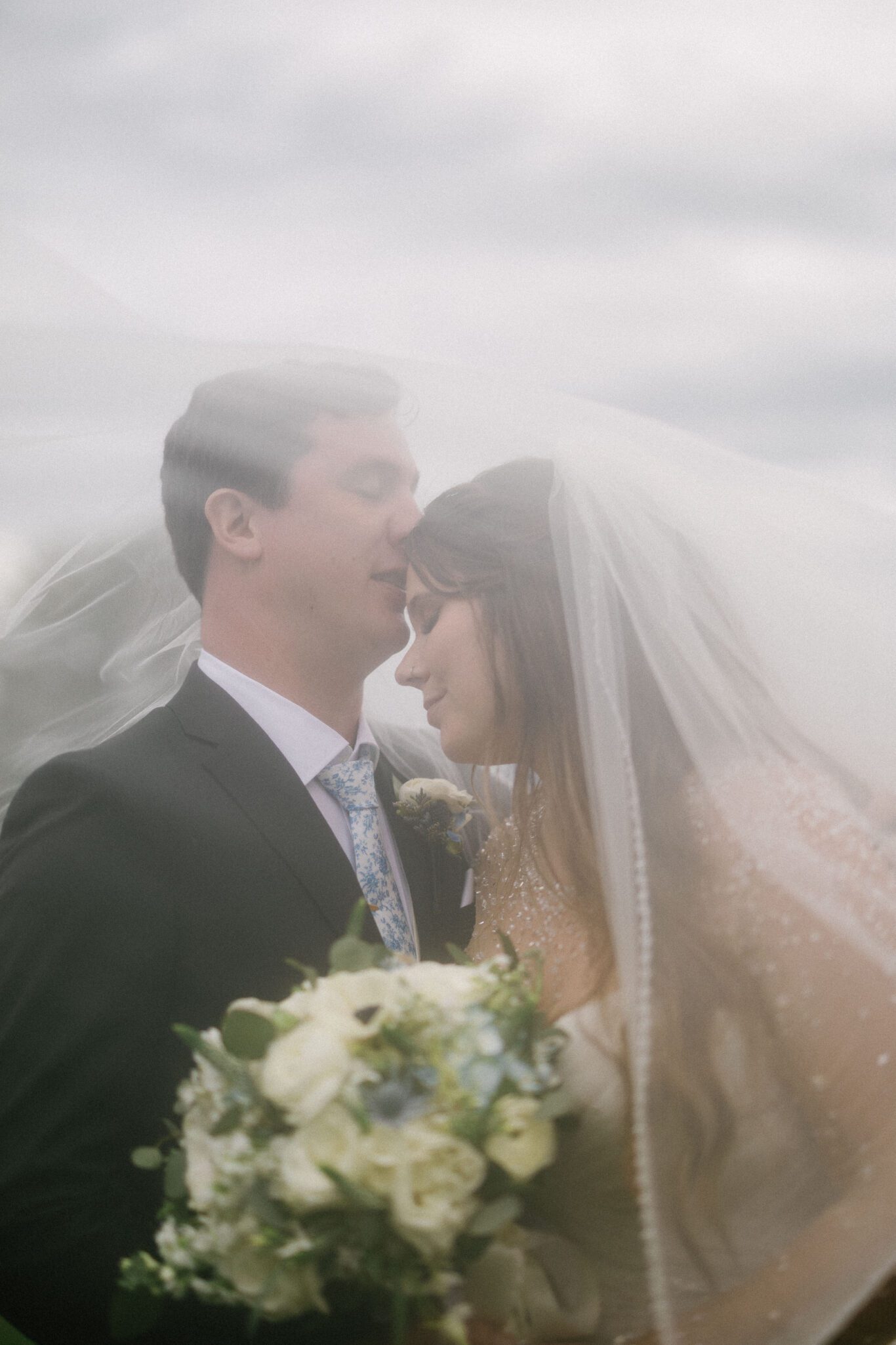 A bride and groom stand close together under a sheer wedding veil. The groom wears a dark suit and floral tie, while the bride holds a bouquet of white flowers and rests her head gently on his chest. The mood is romantic and soft.