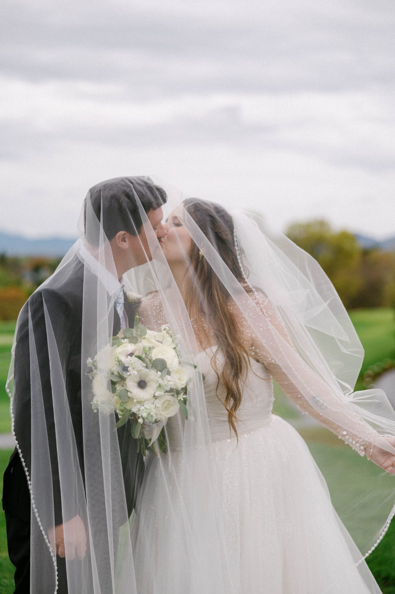 A bride and groom kiss under a sheer veil outdoors. The bride holds a bouquet of white flowers, and both are dressed in wedding attire with a green landscape and cloudy sky in the background.
