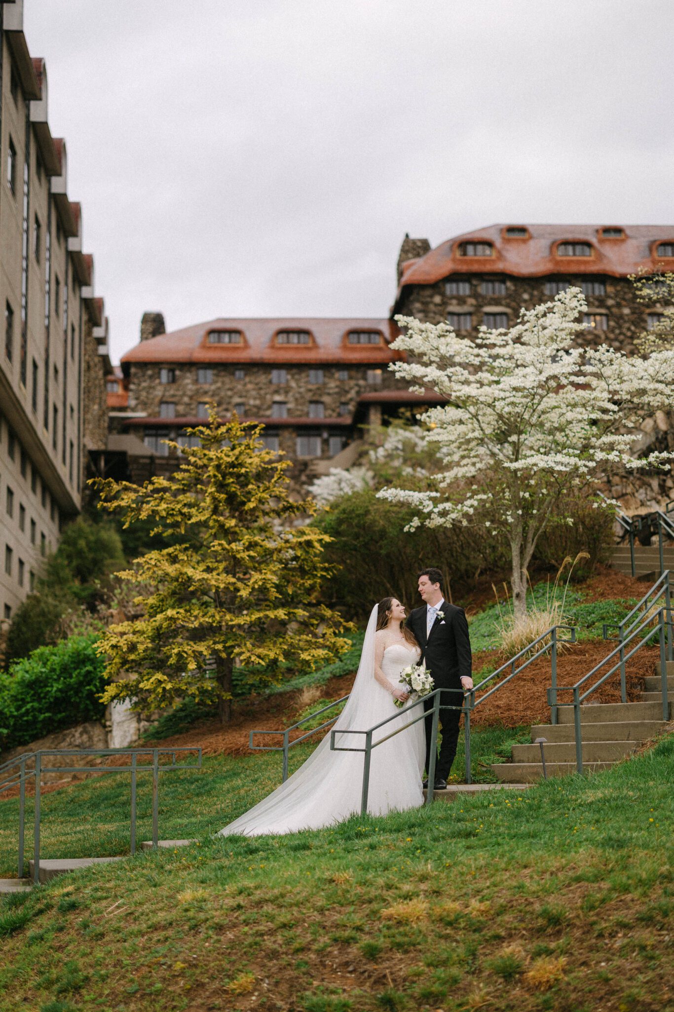 A bride and groom stand on outdoor steps, holding hands and smiling at each other, surrounded by spring trees and greenery, as their Omni Grove Park Inn wedding photographer captures the moment with a stately stone building in the background under a cloudy sky.