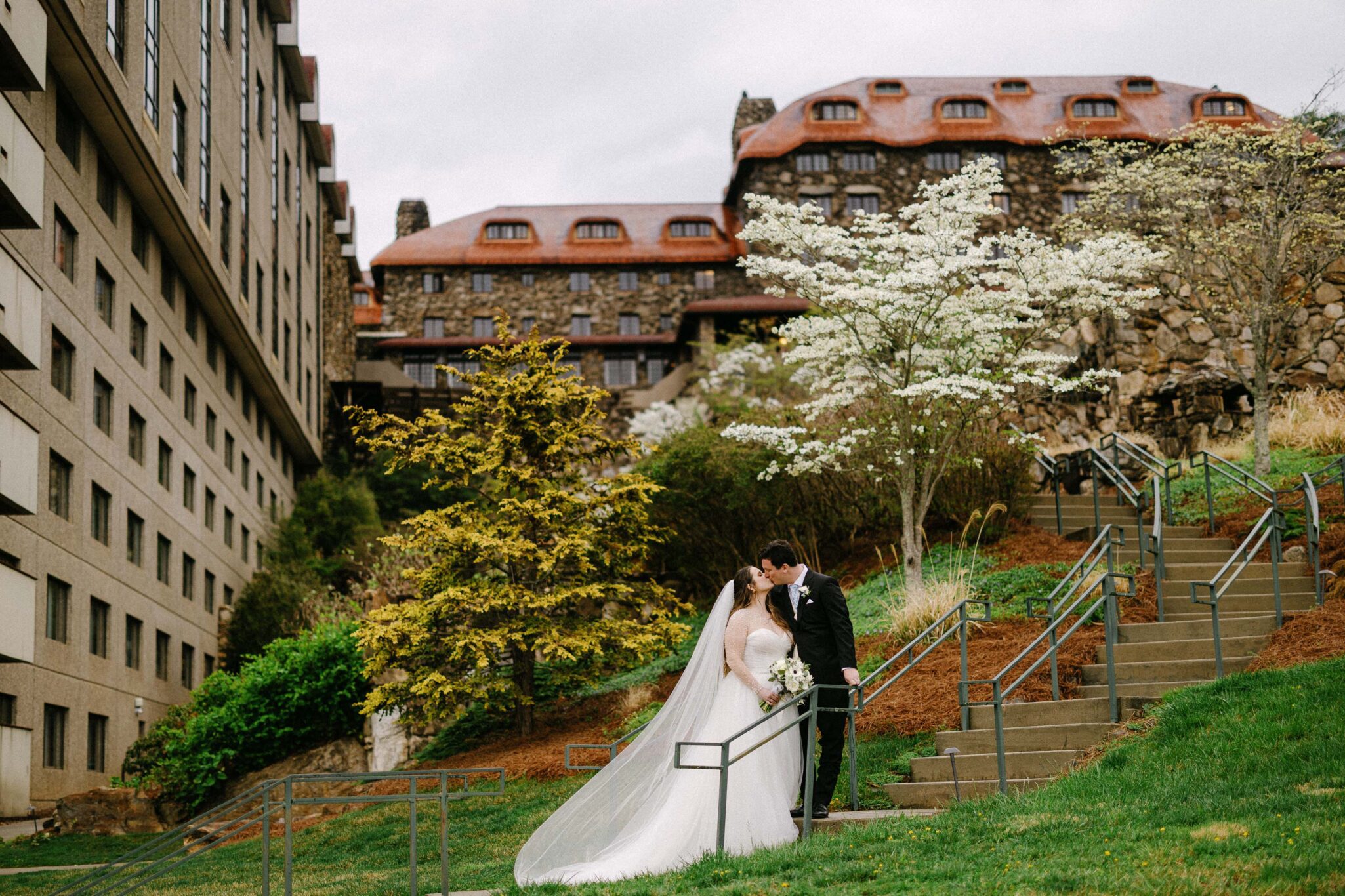 A bride and groom kiss on an outdoor staircase, surrounded by greenery, with historic stone buildings and blooming trees in the background.