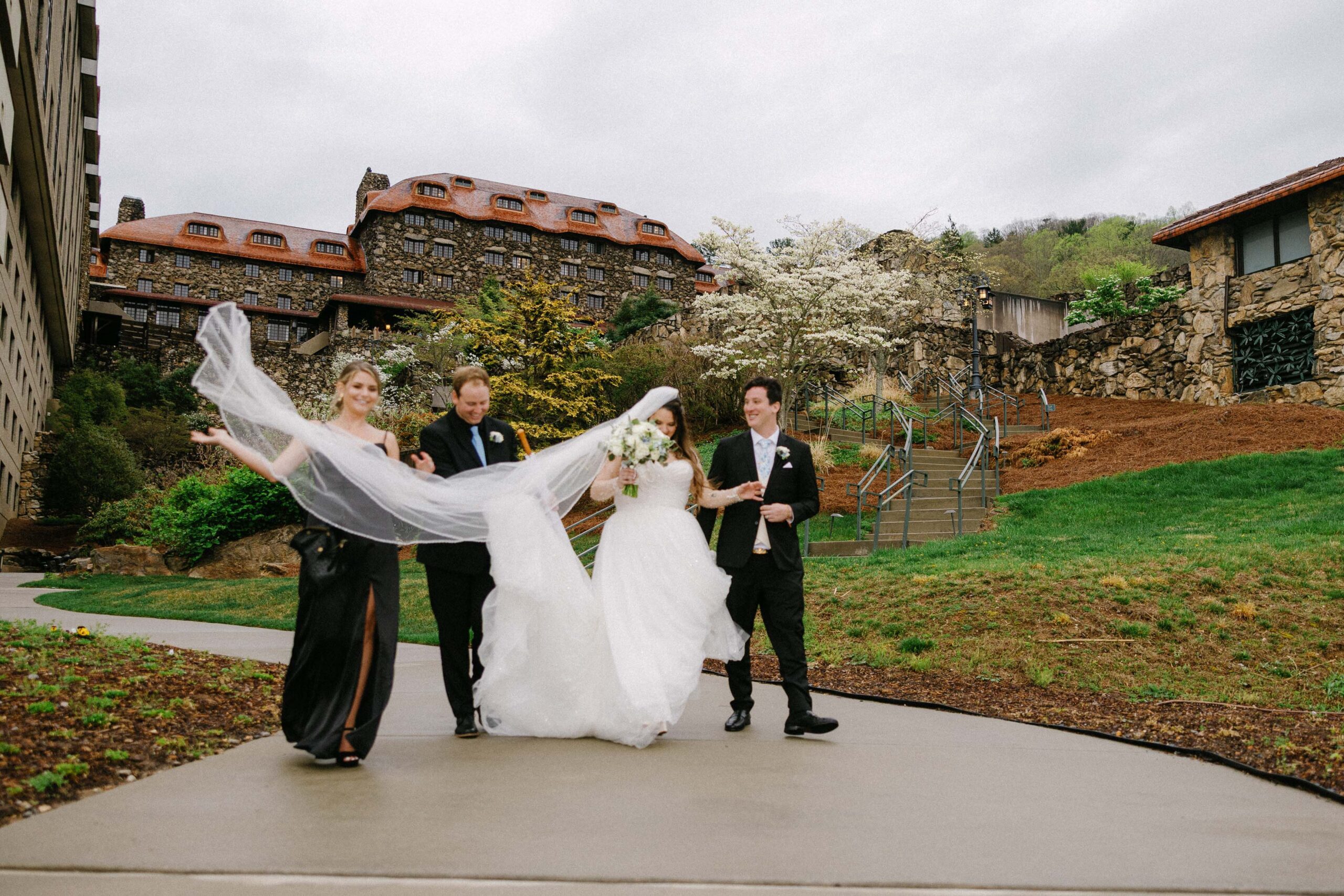 A bride and groom walk outdoors with two attendants. The bride holds a bouquet while her veil is lifted by a bridesmaid. They are near stone buildings and blooming trees on a cloudy day.
