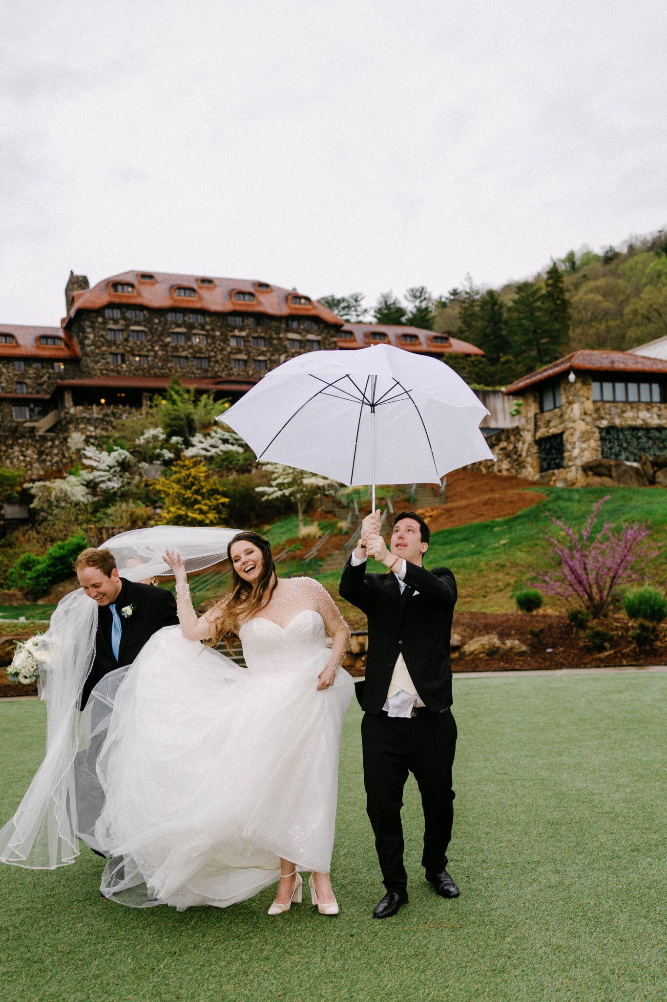 A bride and two groomsmen struggle with wind on a grassy lawn; her dress and veil are blown up while one man holds an inside-out umbrella. Captured by The Omni Grove Park Inn wedding photographer, a grand stone building and trees set the scene.