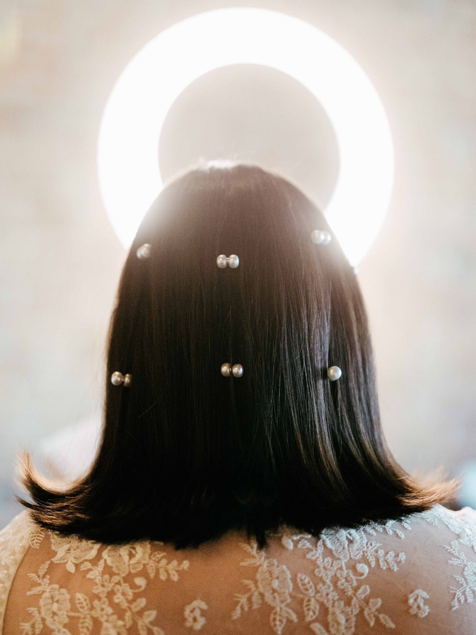 A woman with shoulder-length dark hair, adorned with pearl hairpins, is seen from behind. She wears a lace garment, and a bright circular light behind her head creates a halo effect.