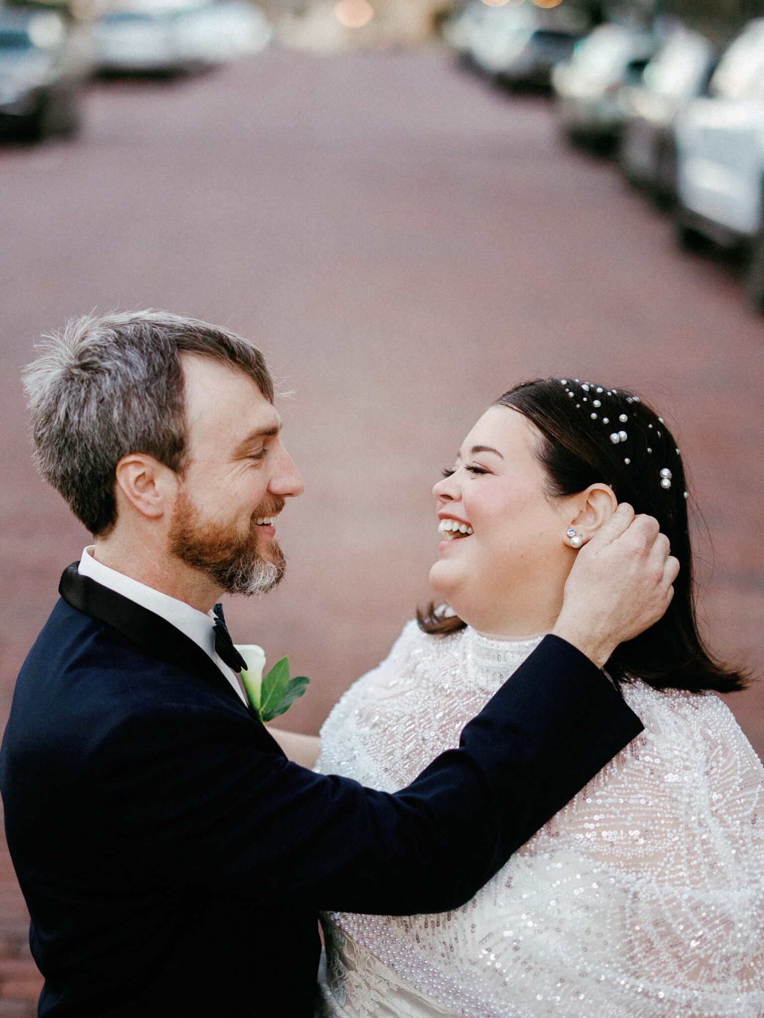 A groom in a suit gently holds his smiling bride, who wears a beaded white dress and pearls in her hair, as they share a joyful moment on a brick-paved street lined with cars.