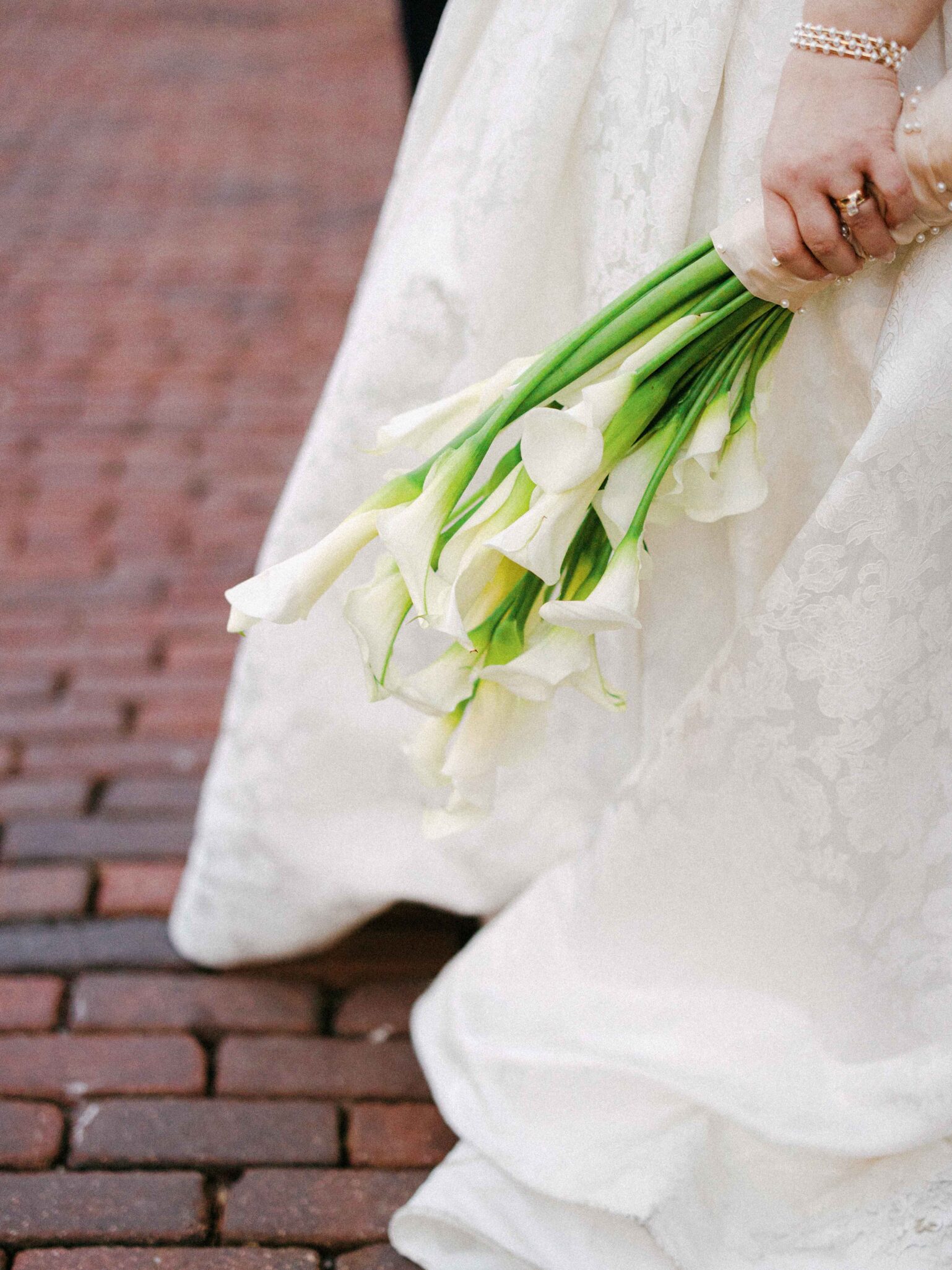 A bride in a white lace dress holds a bouquet of white calla lilies. She stands on a red brick path at The Venue in downtown Asheville, her hand adorned with pearl jewelry and a ring—perfect for timeless newlywed portraits.