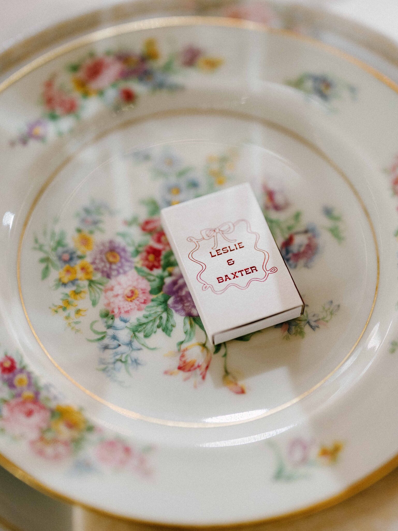 A small matchbox labeled Leslie & Baxter rests on a decorative floral china plate with gold trim.