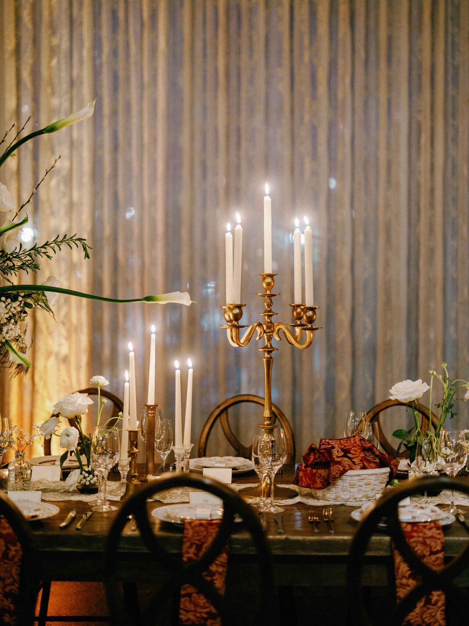 Elegant dining table set for a formal event with gold candelabras holding lit candles, white flowers, glassware, and decorative napkins, against a shimmery curtain backdrop.