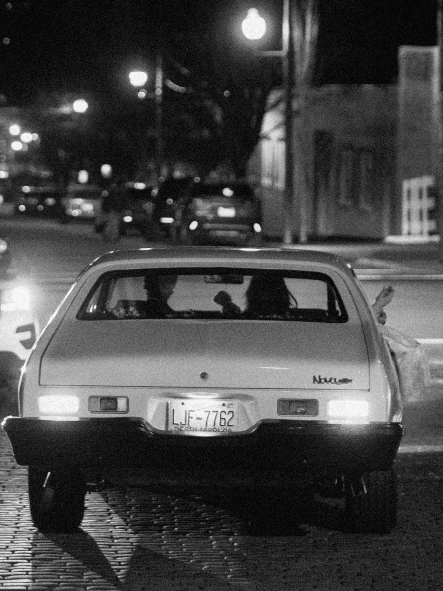 A black and white wedding send off: a car drives at night on a cobblestone street outside The Venue Downtown Asheville, with two people inside. The glowing streetlights illuminate the city as their journey begins.