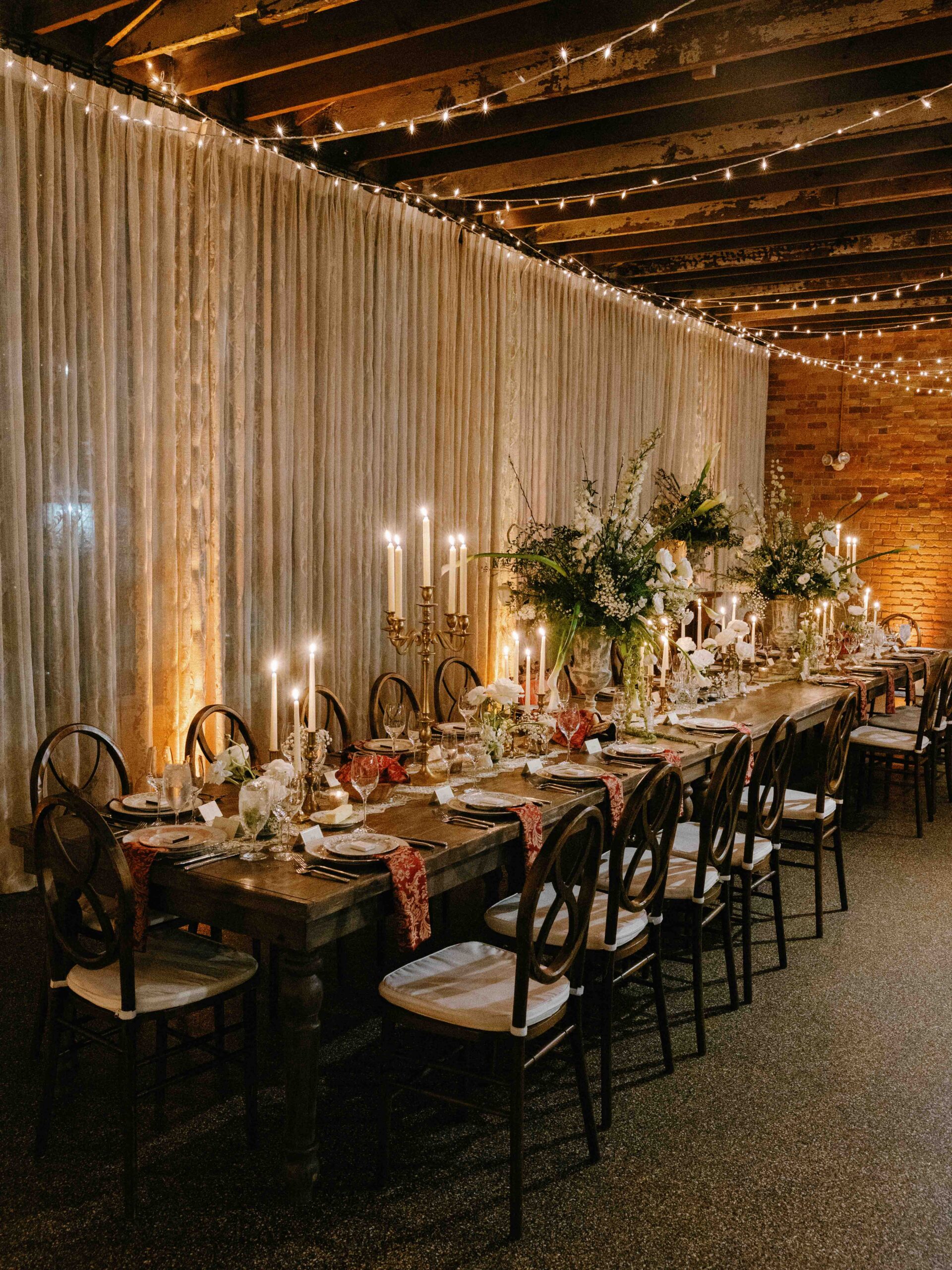 A long, rustic dining table at The Venue Downtown Asheville Weddings is elegantly set with candles, floral centerpieces, and arranged plates. Wooden chairs surround the table beneath string lights and sheer curtains against exposed brick walls.