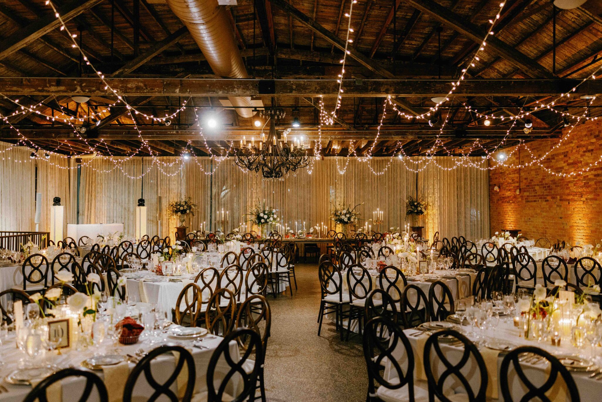 Elegant wedding reception hall at The Venue Downtown Asheville Weddings, featuring round tables set with white linens, floral centerpieces, candles, and string lights hanging from exposed wooden beams. Warm and inviting atmosphere.