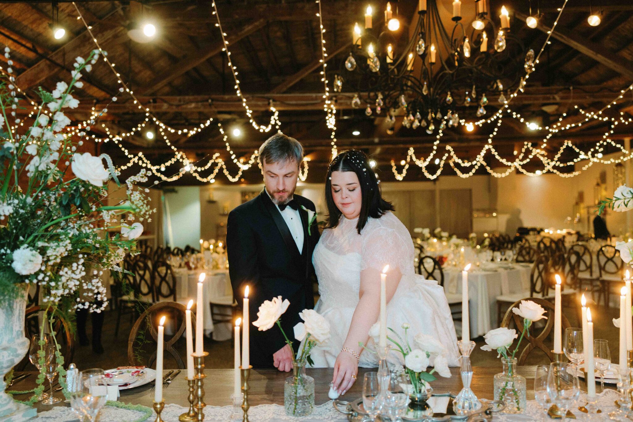 A bride and groom in formal attire stand together at a decorated table with white flowers and candles, under string lights and a chandelier in a rustic, warmly lit wedding venue.