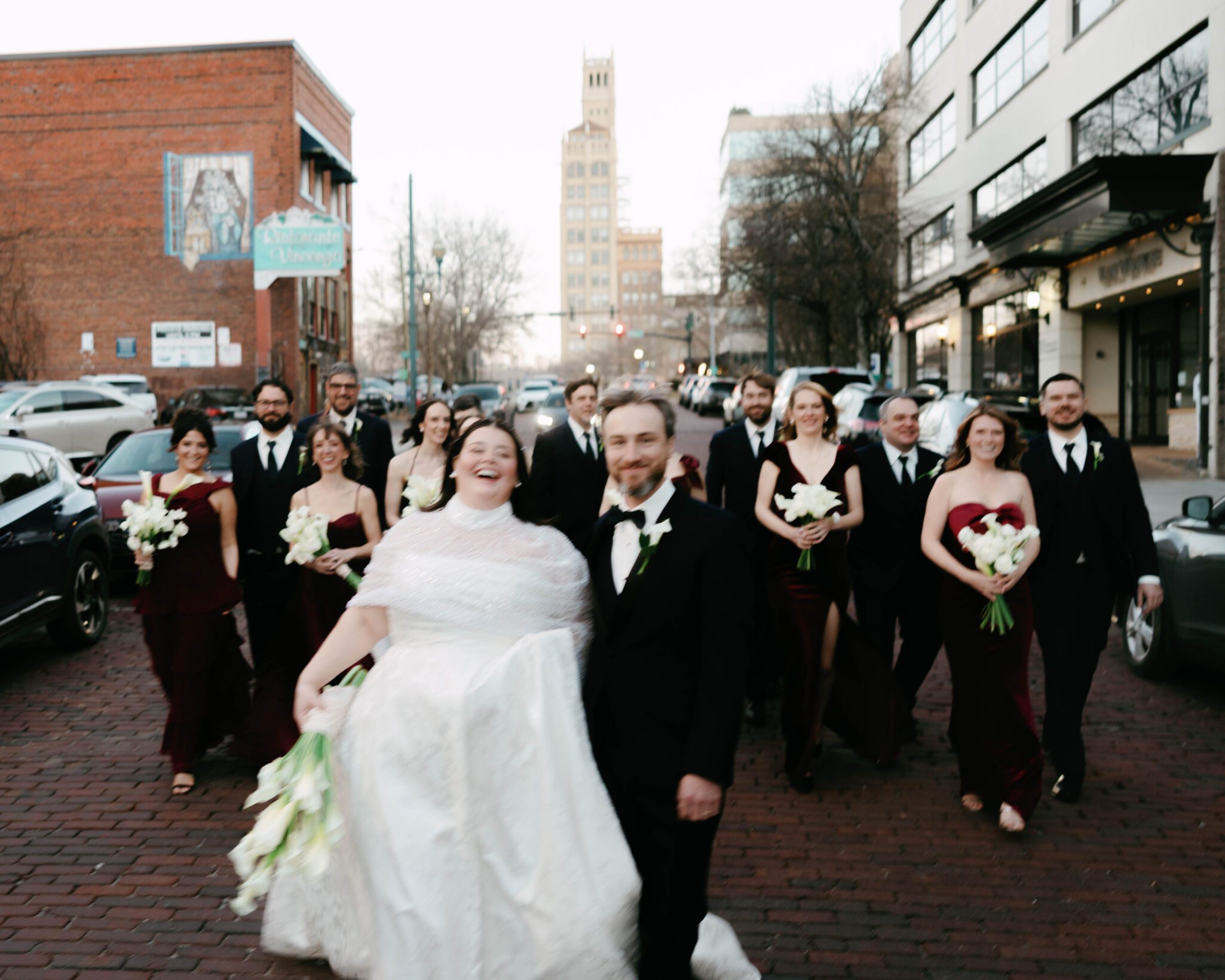 A joyful bride and groom, Leslie + Baxter, walk arm in arm down a brick street after their Downtown Asheville Wedding, followed by their wedding party in formal attire, with buildings and cars lining the street in the background.